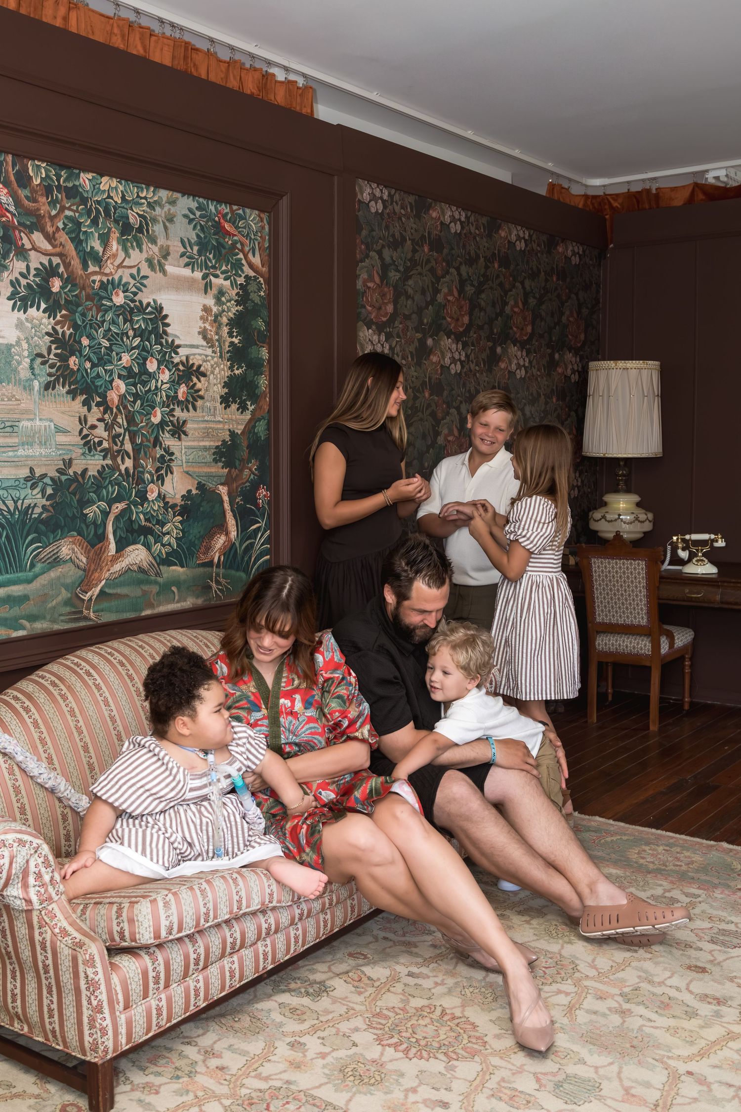 Multi-generational family gathering in elegant living room with tapestry walls, children on sofa with parents nearby.