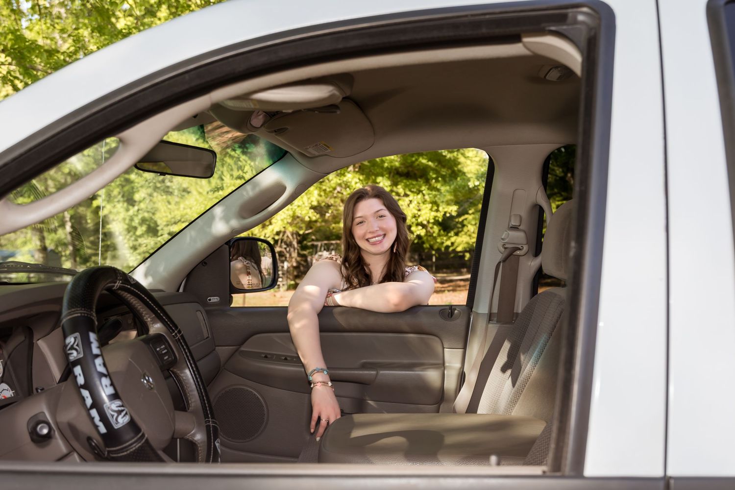 Smiling young woman sitting in driver seat of white truck, leaning out open window on sunny day.