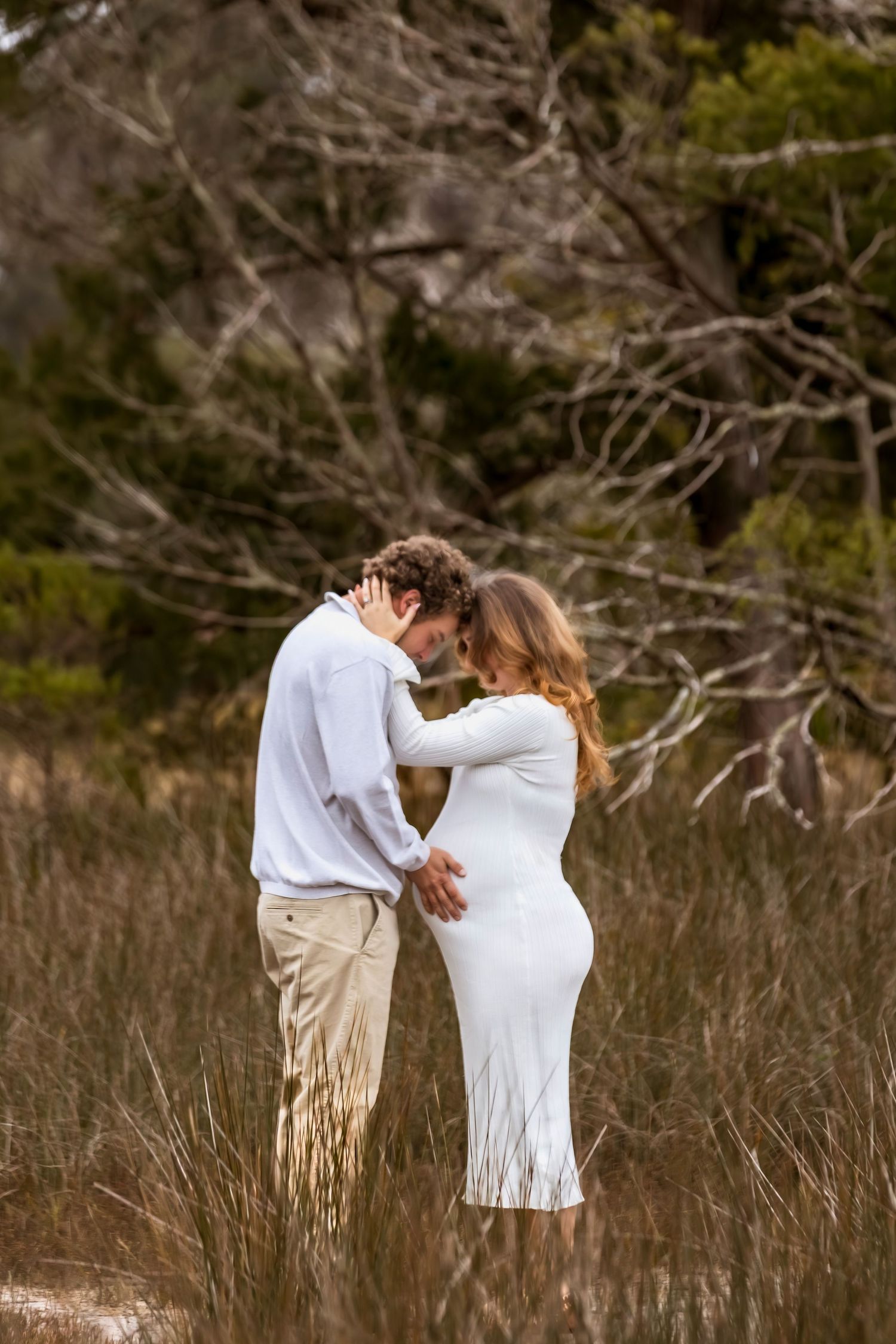 A romantic couple embraces in a field of tall grass during golden hour with bare branches in the background.