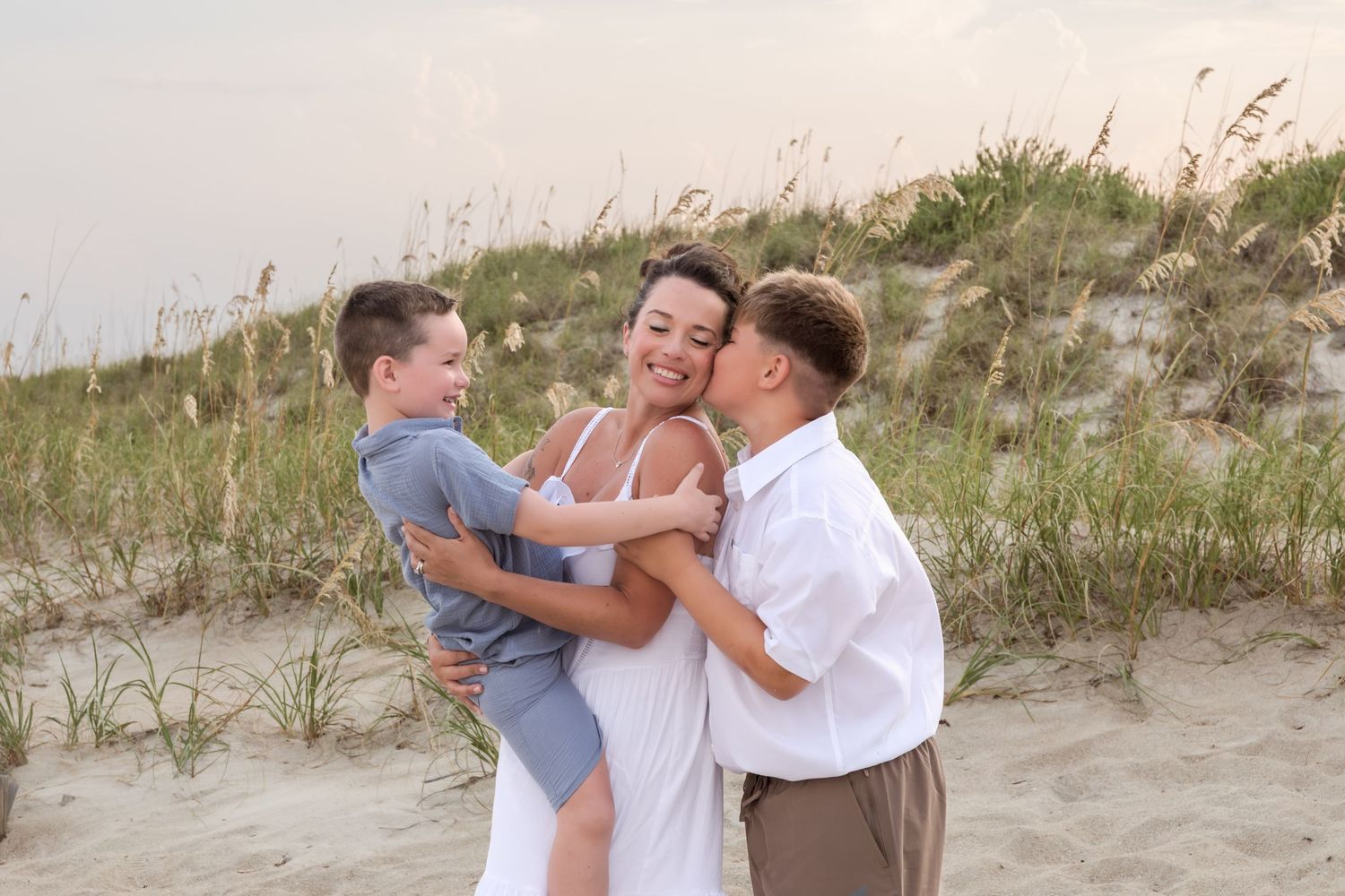 A mother smiles joyfully as her two young sons hug her on a sandy beach with sea oats in the background.