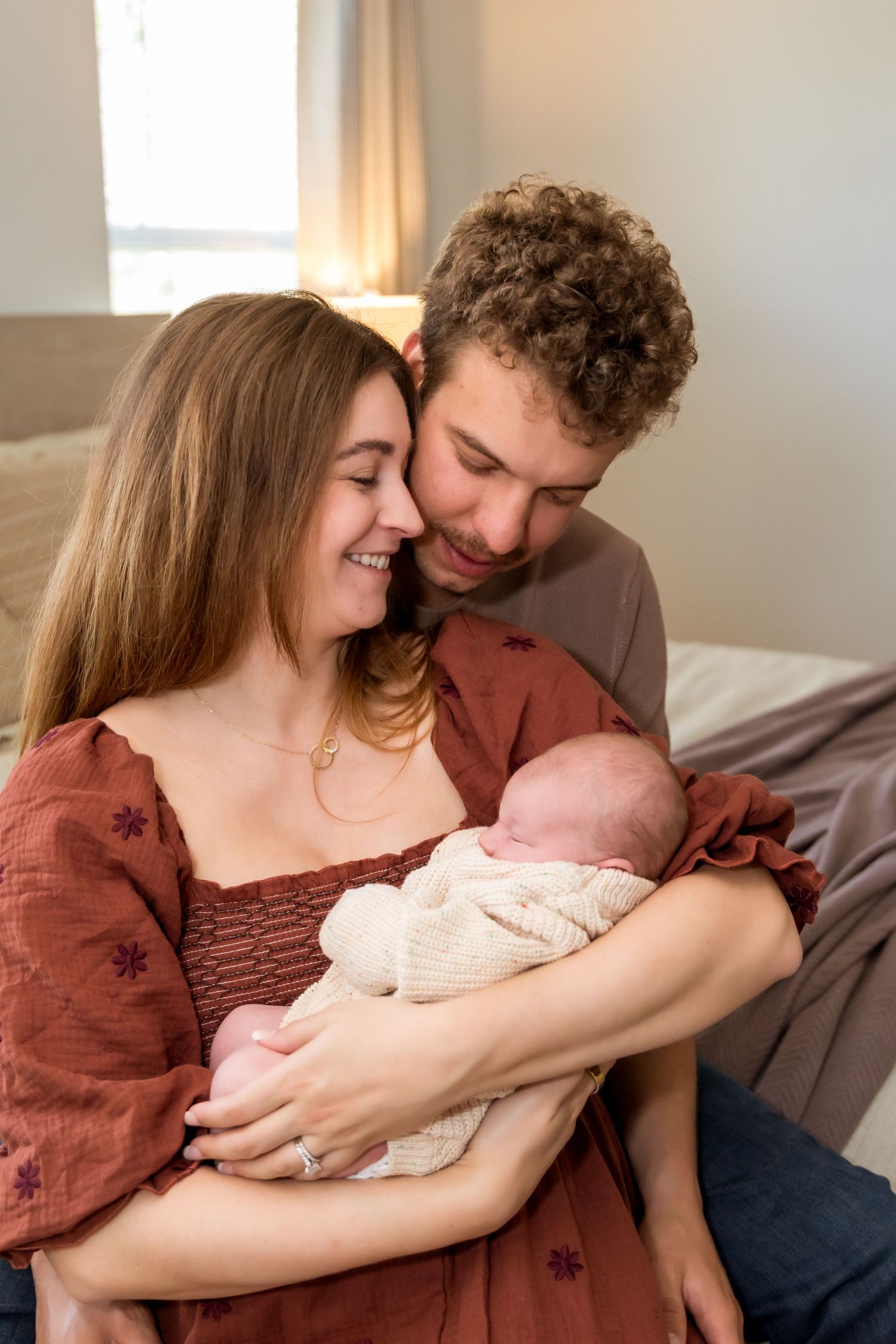 Smiling young couple share a joyful moment holding their newborn baby wrapped in white knit blanket.
