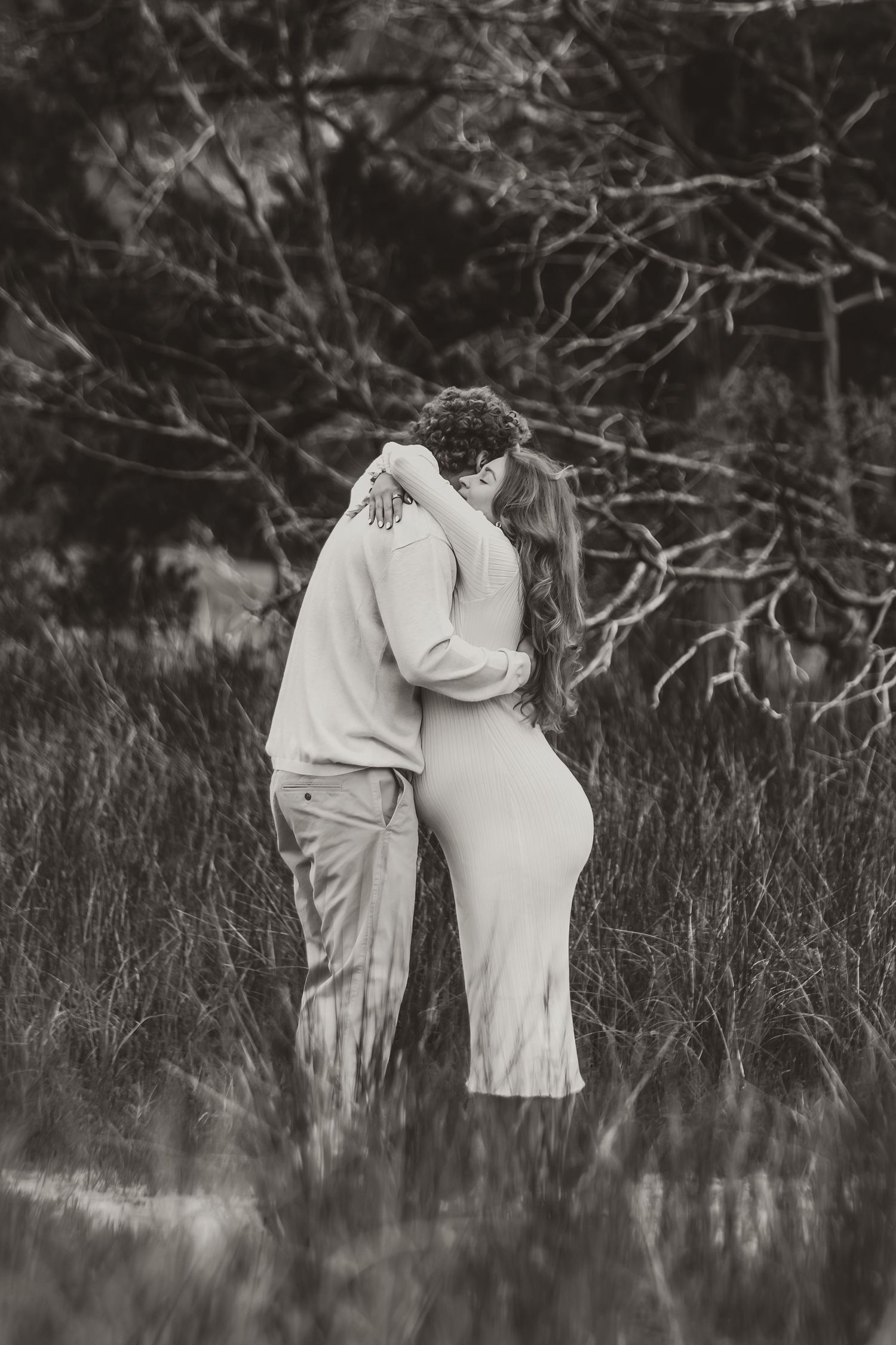A couple embraces intimately in black and white photography against a natural outdoor backdrop with bare branches.