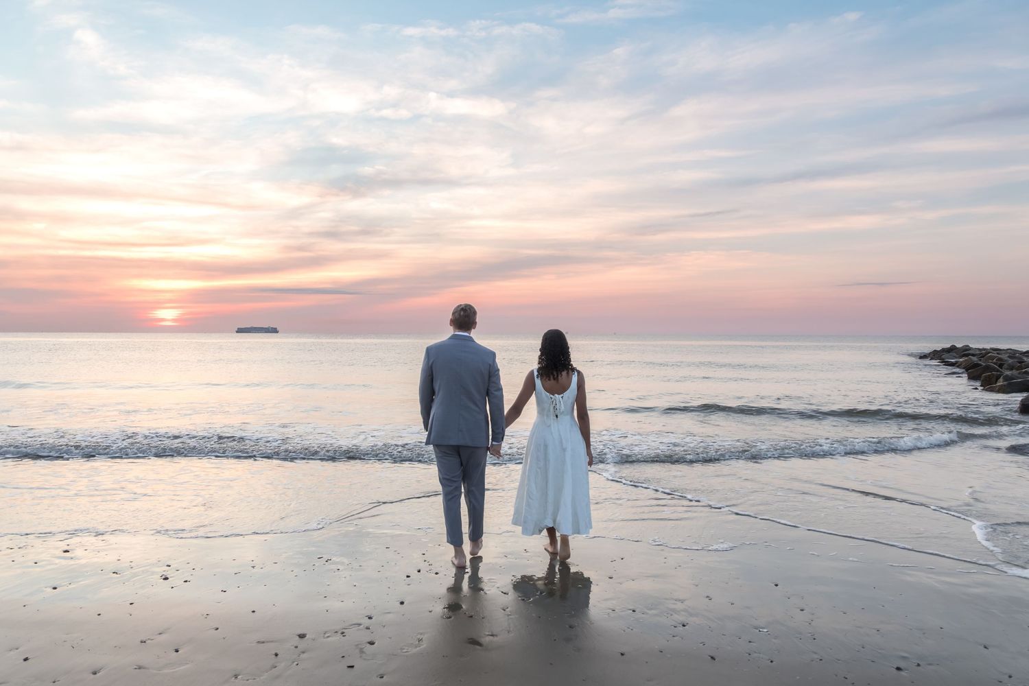 Couple holding hands on beach at sunset, man in gray suit and woman in white dress facing the ocean.