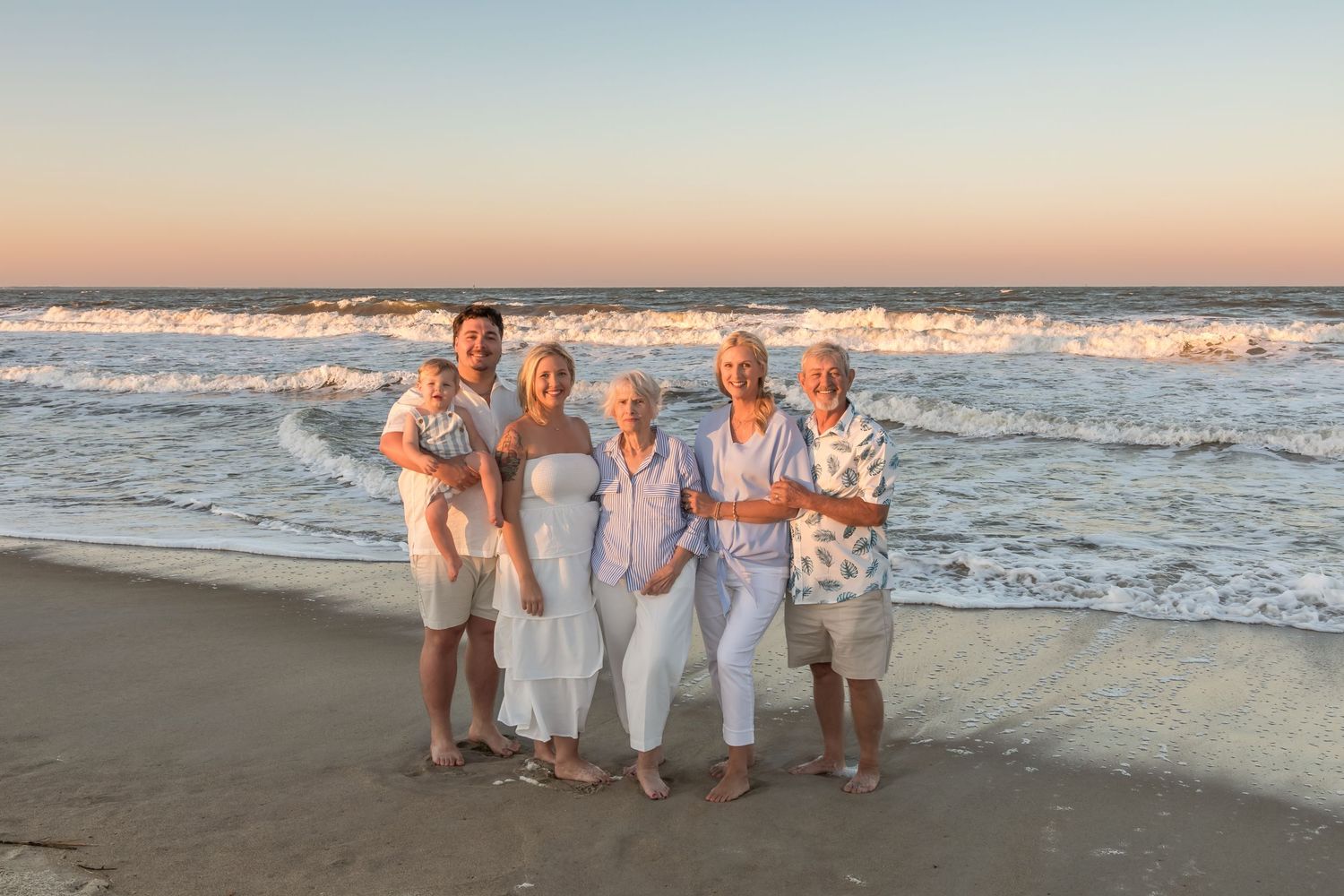 Multi-generational family smiling together on a sandy beach at sunset with ocean waves in the background.