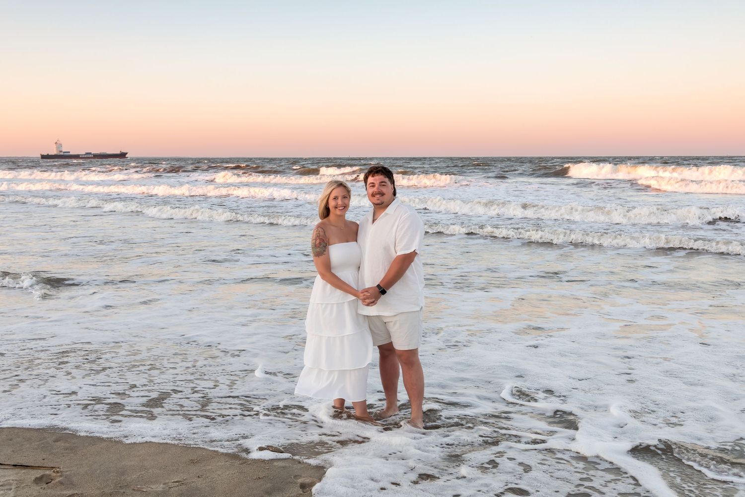 Pregnant couple in white outfits embracing on a sandy beach at sunset with ocean waves behind them.
