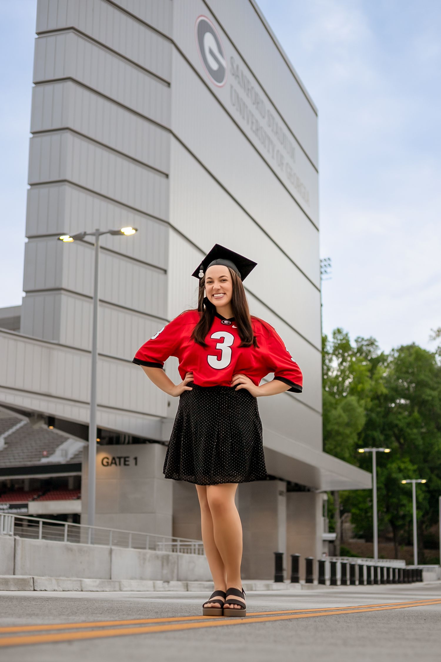 Graduate in red jersey and cap poses confidently outside stadium Gate 1.