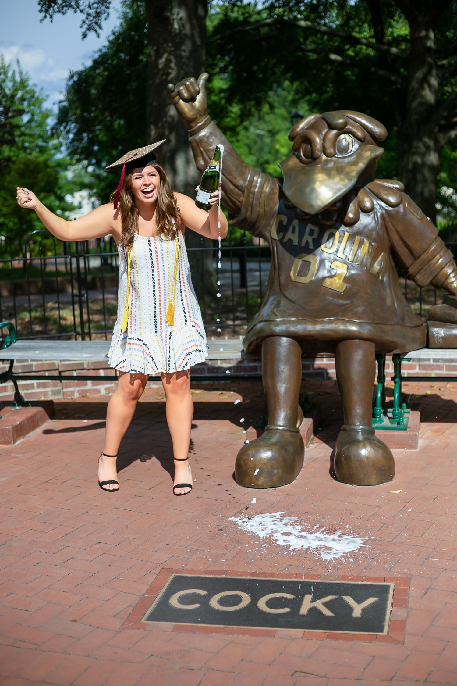 Graduate posing playfully with bronze statue on brick courtyard.