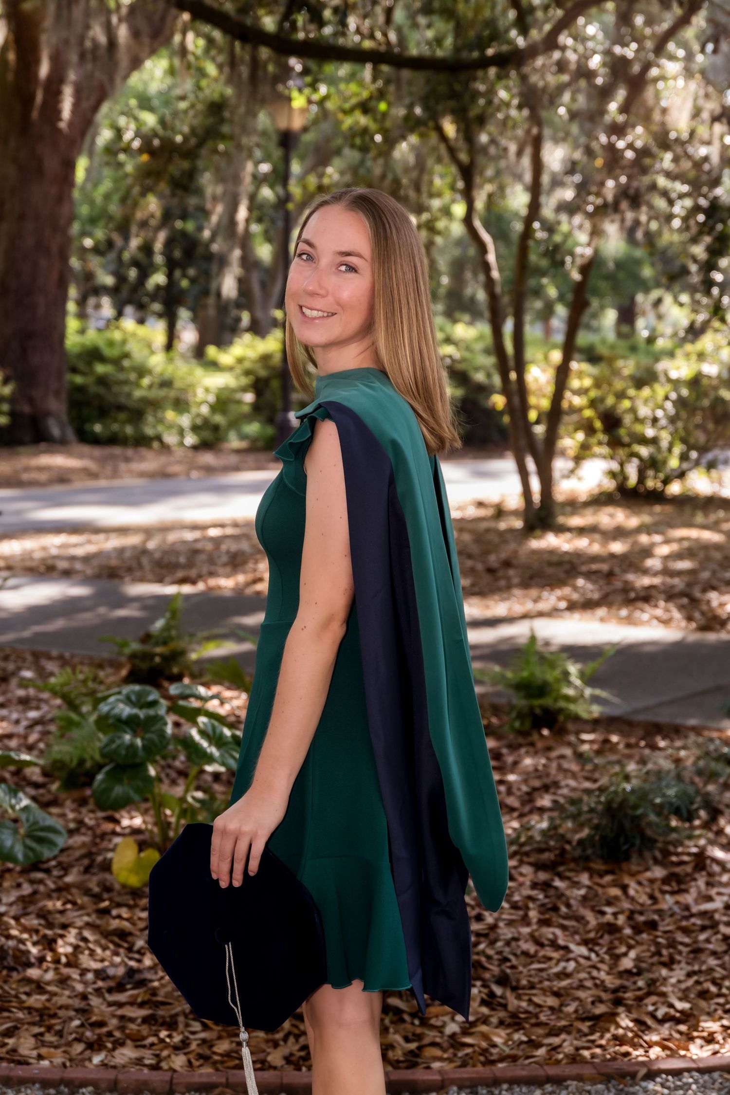 Young woman in green graduation gown smiling over shoulder, holding cap in tree-lined park setting.