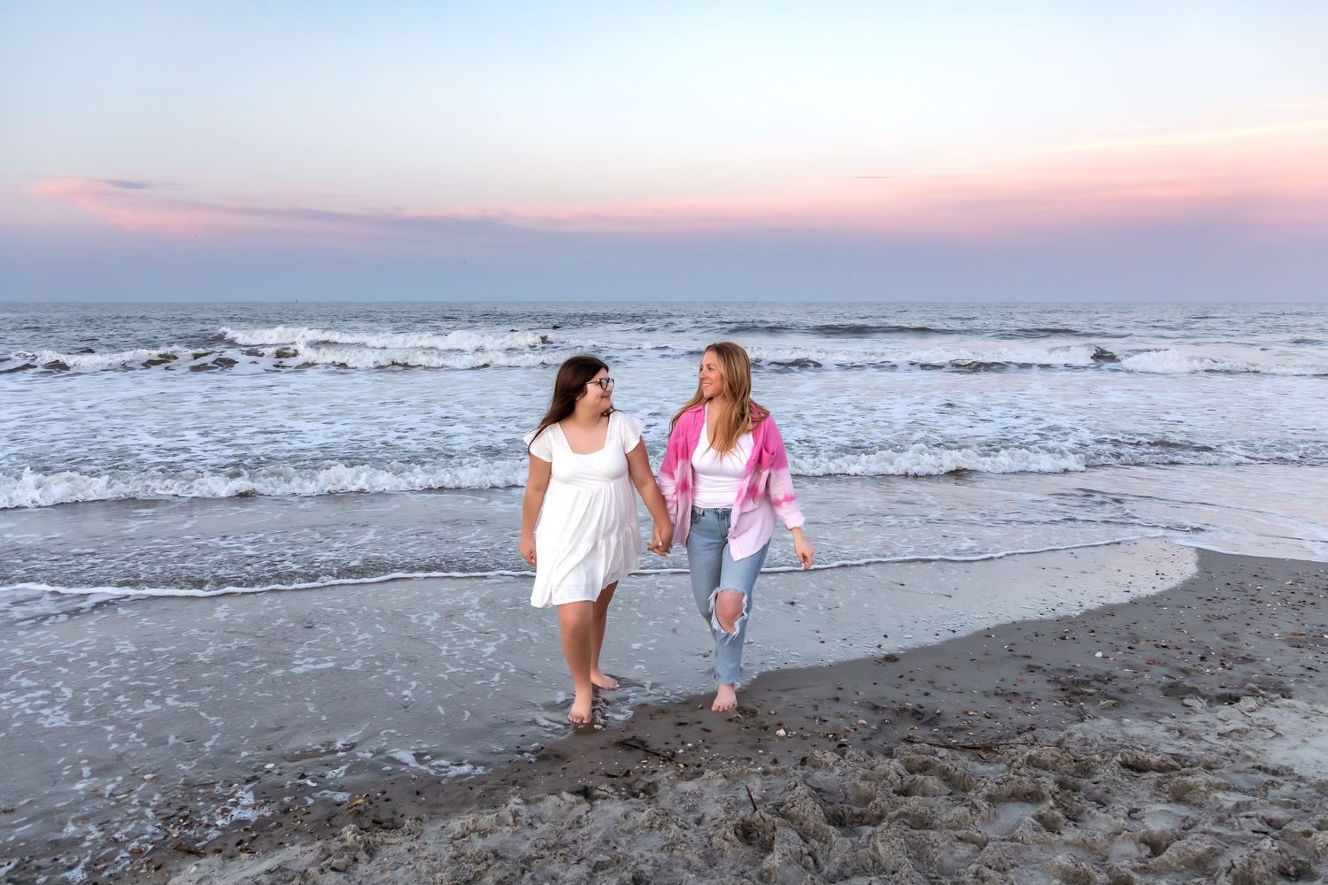 Two women holding hands and walking barefoot on a sandy beach at sunset with ocean waves in the background.