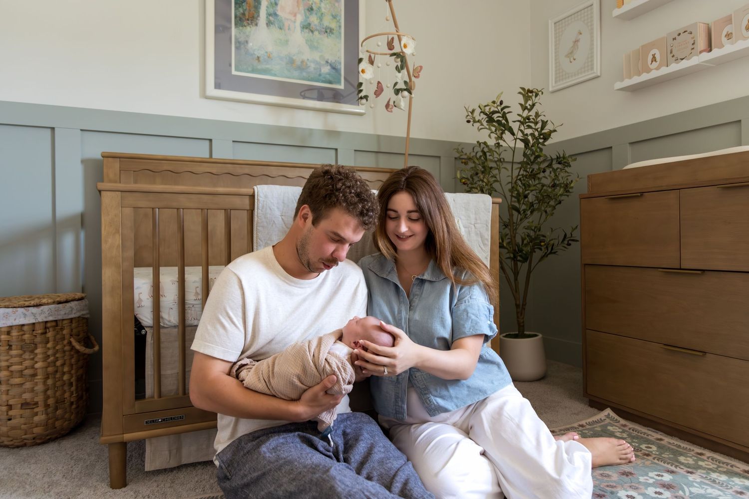 Young parents sit on nursery floor admiring their swaddled newborn baby together.