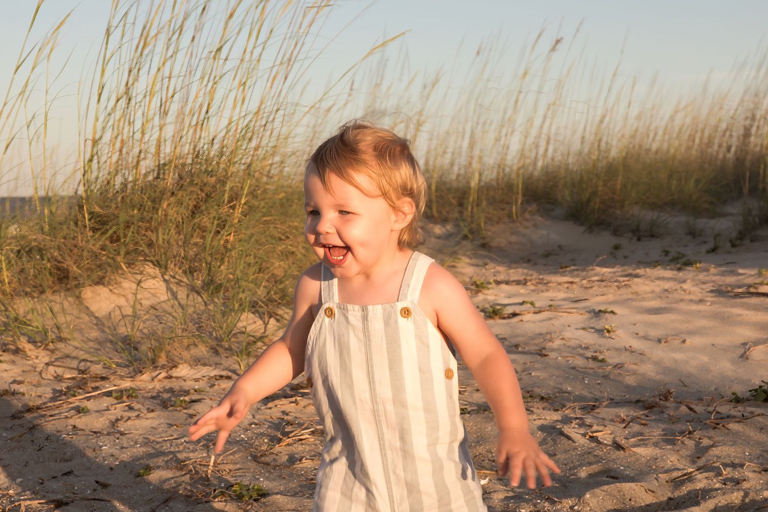 Happy toddler in striped overalls laughing and running on a sandy beach with tall grass dunes in the background.