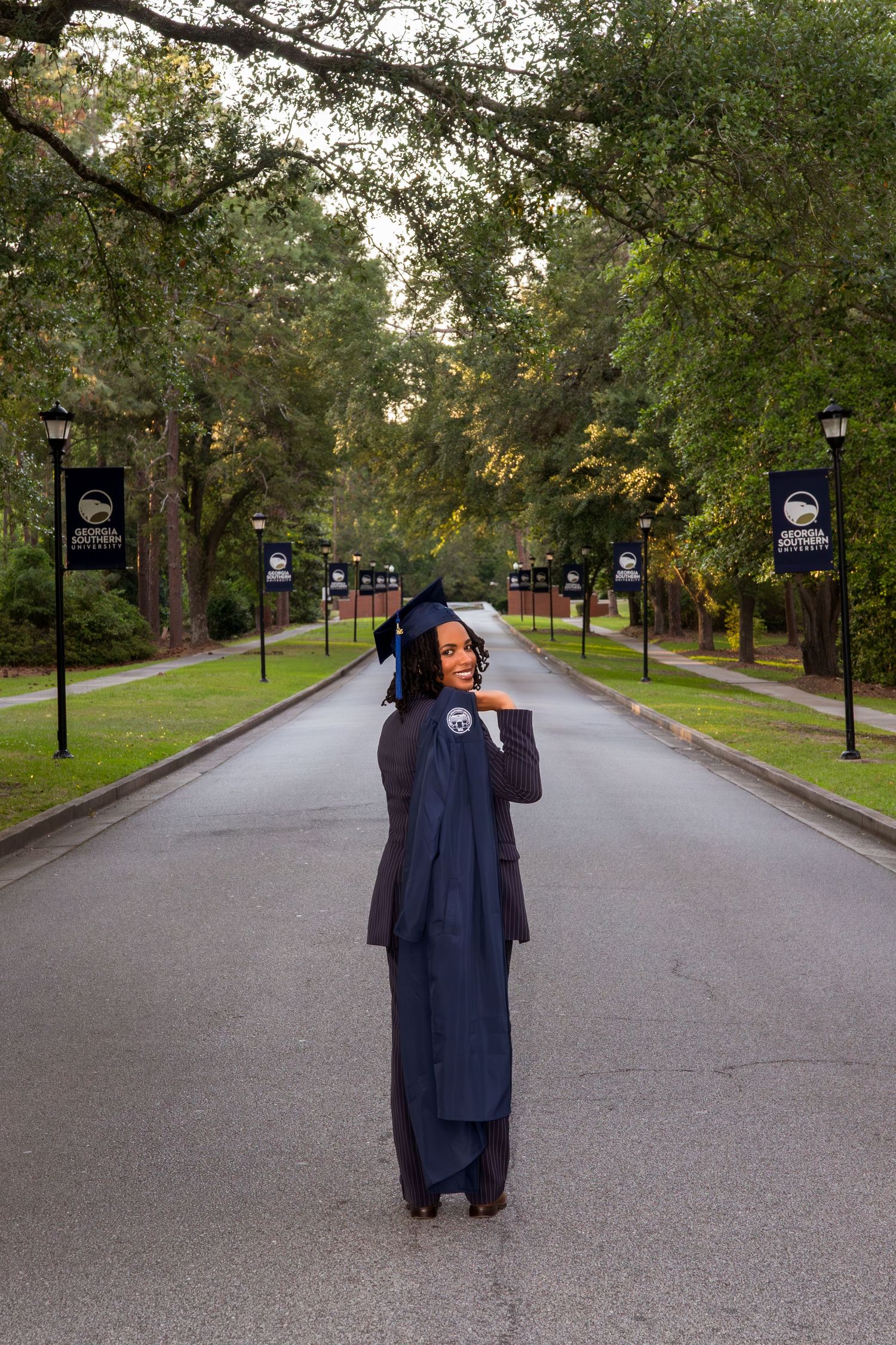 Graduate in navy blue cap and gown walking down a tree-lined campus path.
