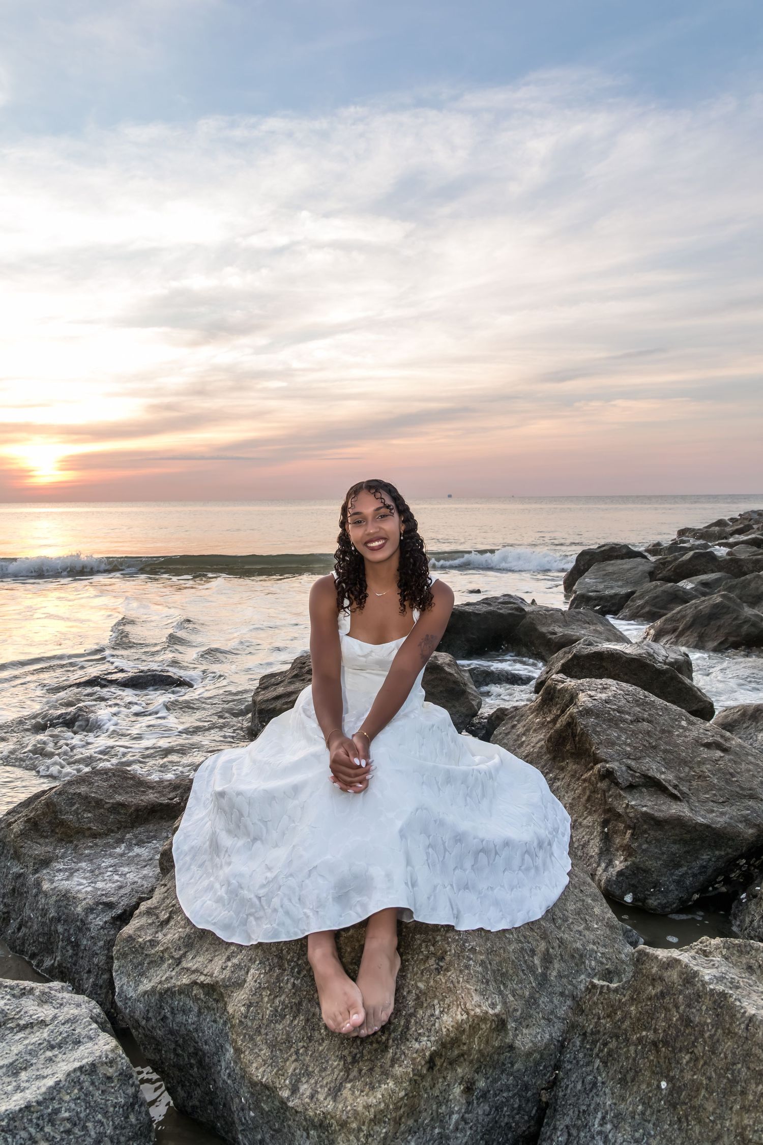 Young woman in white dress sits on rocks at sunset beach, smiling with ocean waves behind her.
