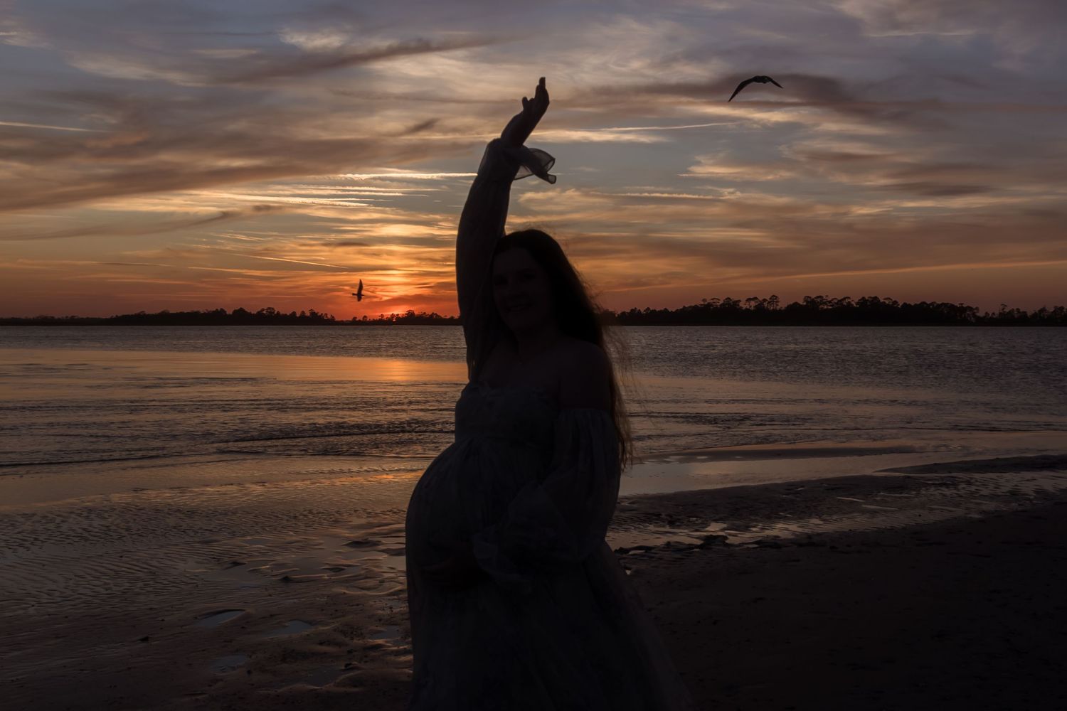 Silhouette of a woman dancing on a beach at sunset with vibrant orange sky and calm water in the background.