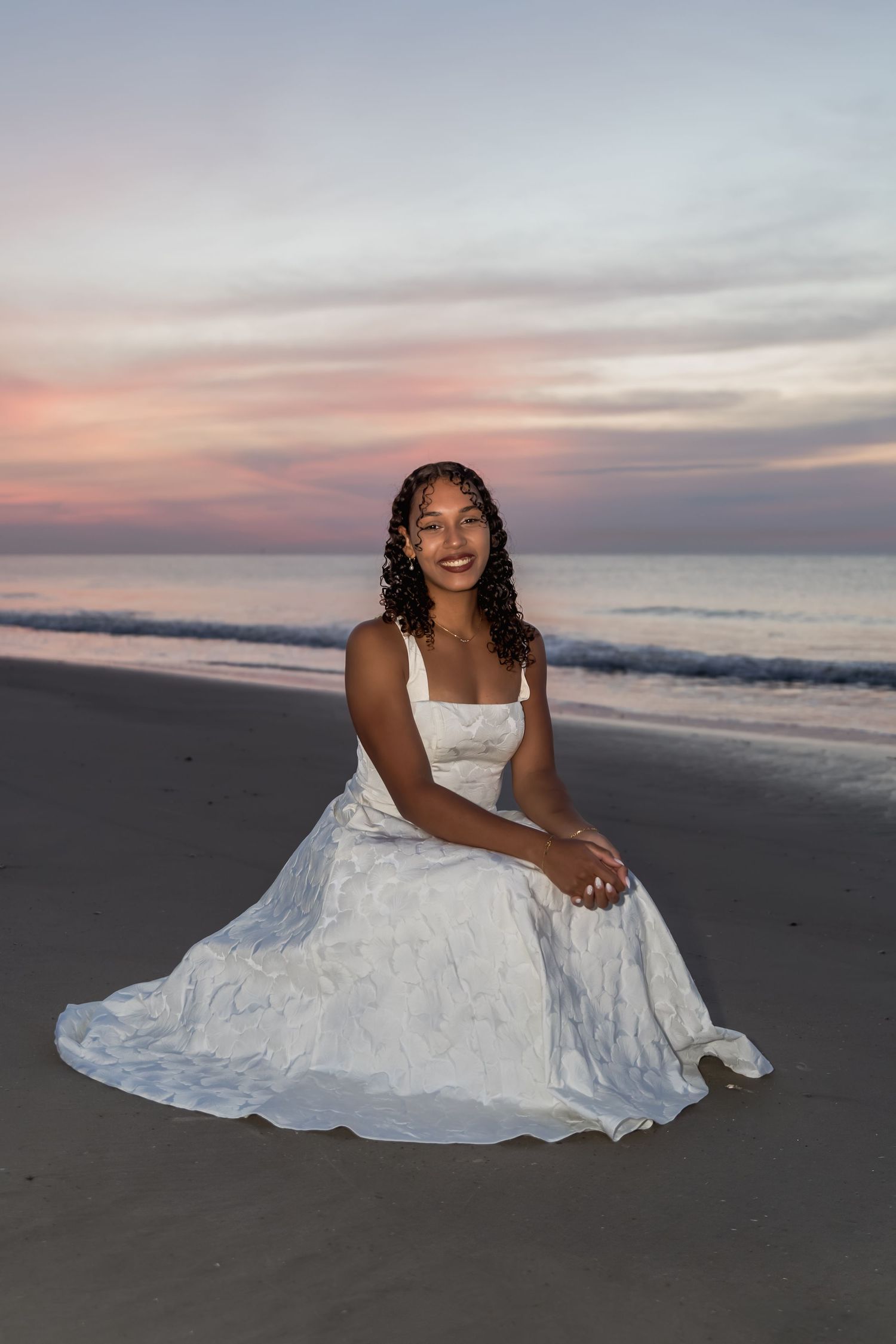 A smiling bride in a white gown sits on a sandy beach at sunset with ocean waves behind her.