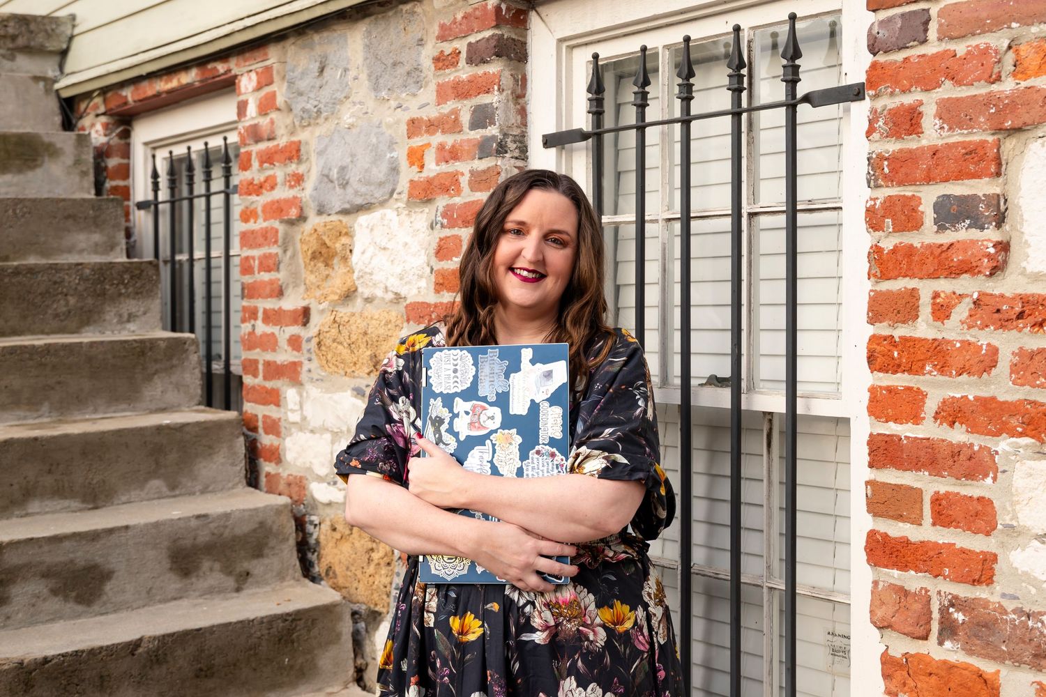 Woman in floral dress poses near red brick wall with black iron window bars.