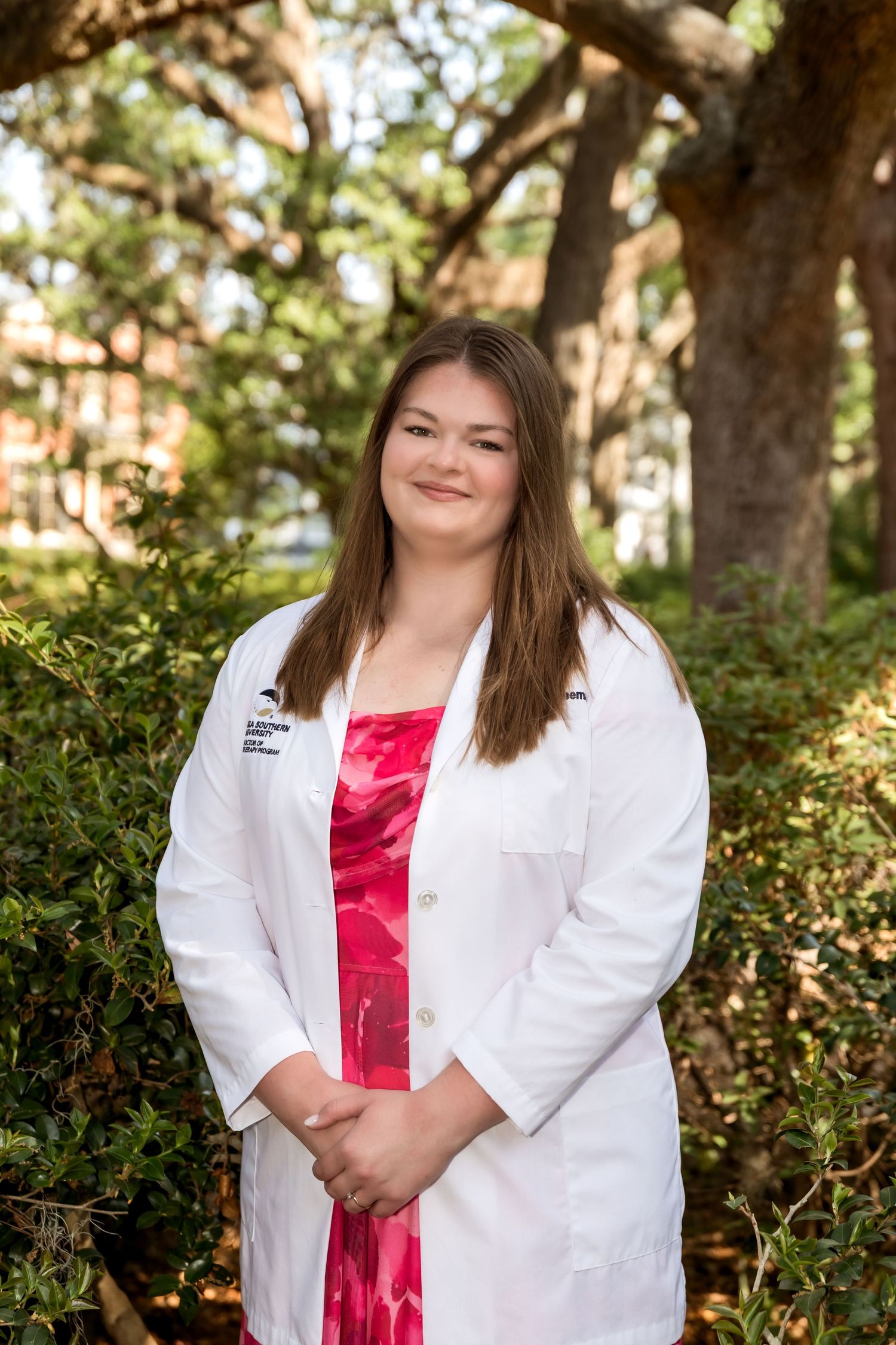 Young woman in white lab coat and pink dress smiles outdoors under oak trees.