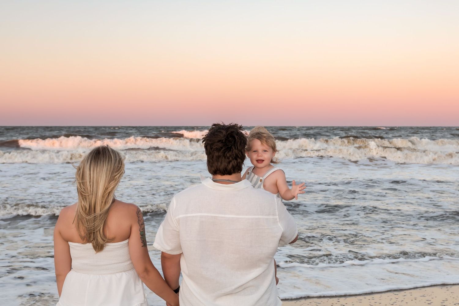 Family of three enjoying a beautiful sunset at the beach, parents holding baby girl while waves crash behind them.