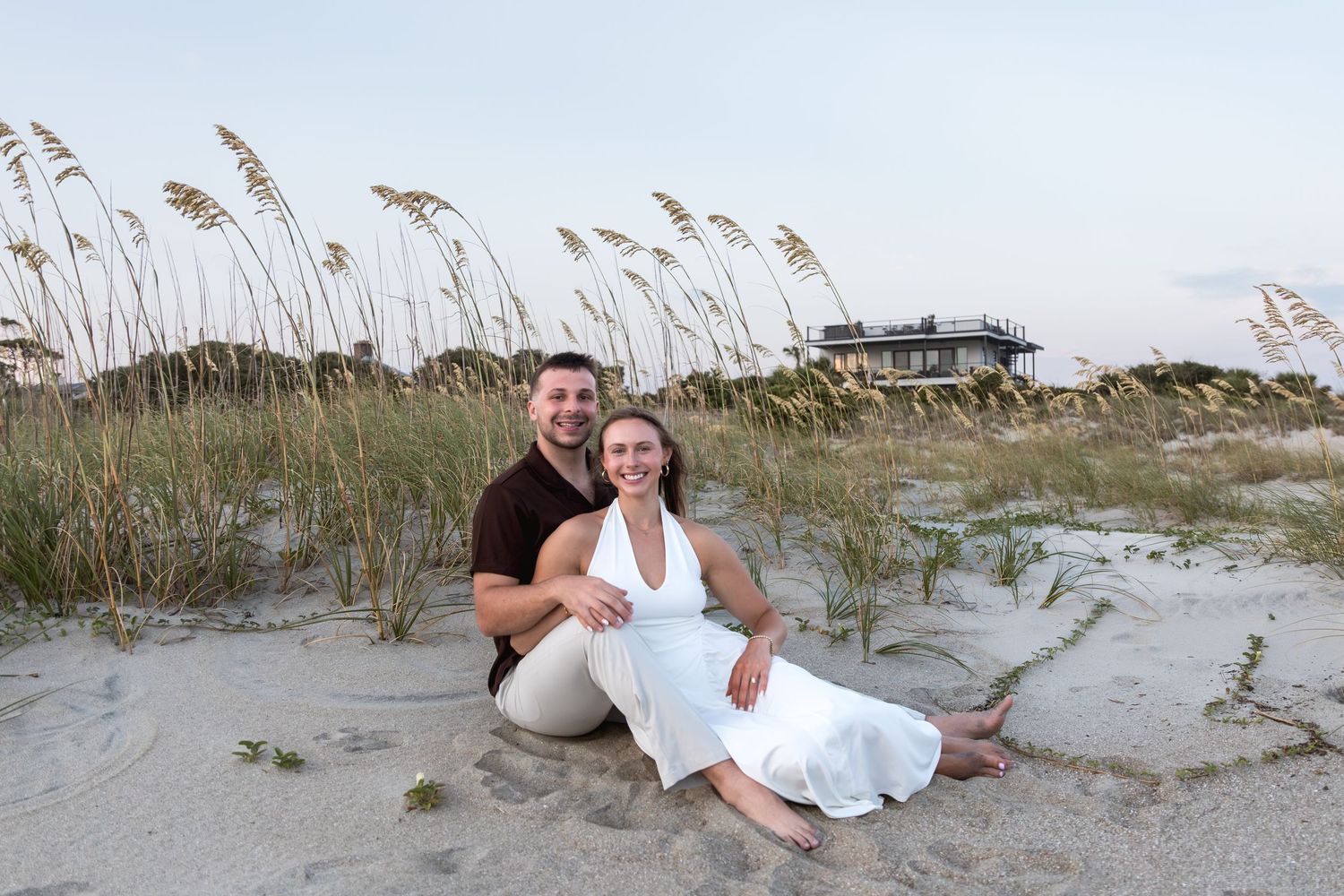 Couple sitting together on sandy beach with sea oats and coastal house in background at dusk.