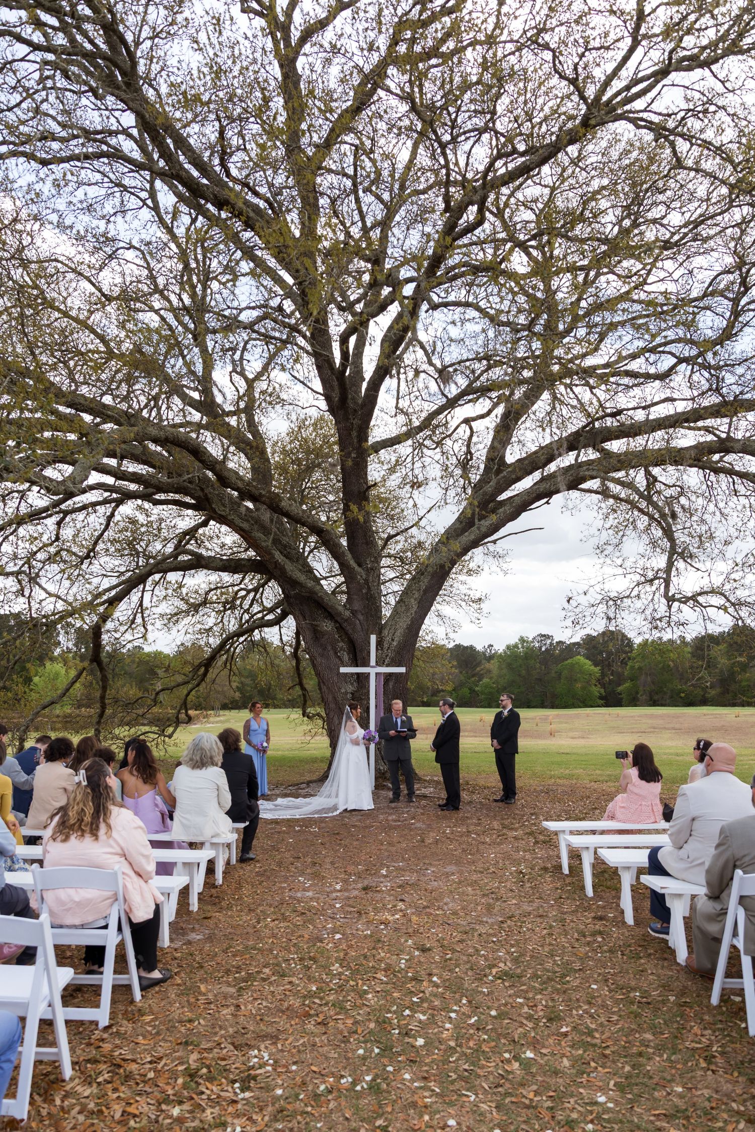 Outdoor wedding ceremony under a large oak tree with white chairs, a cross, and guests gathered on a leaf-covered ground.