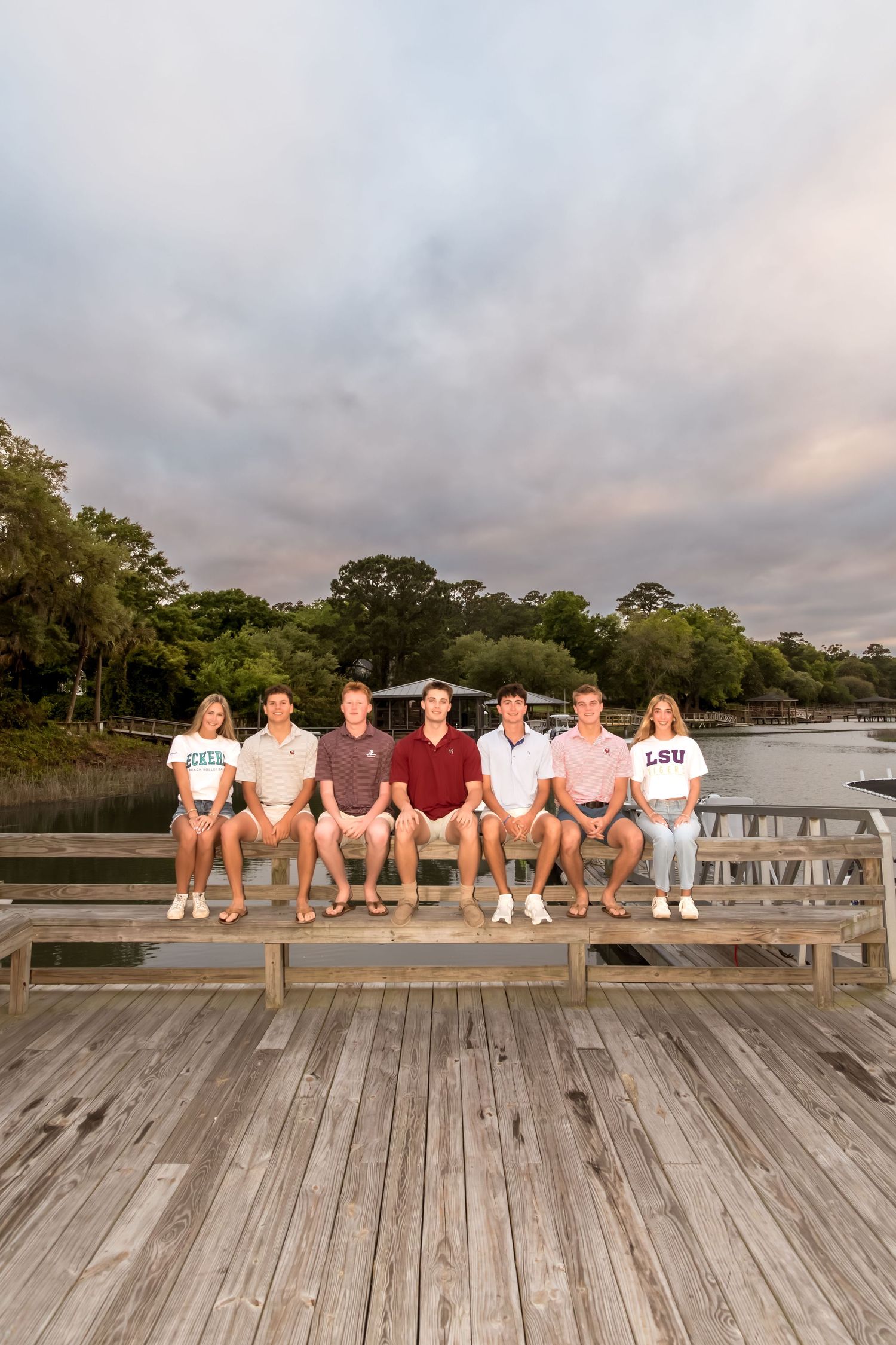 Seven young adults sit together on a wooden dock near a serene waterway at dusk, wearing college shirts including LSU.