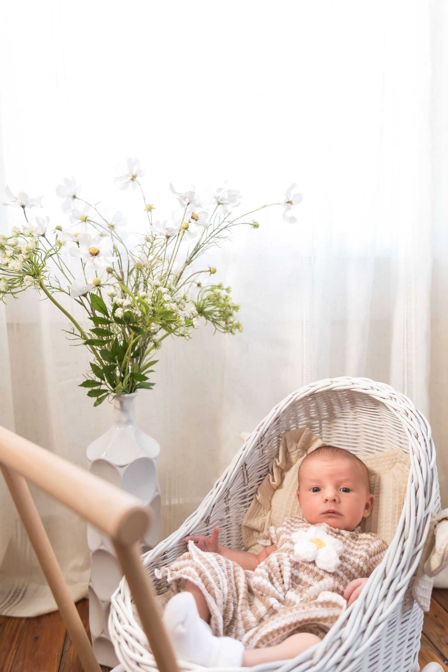 Newborn baby lying in a white wicker bassinet beside a vase of white wildflowers near a bright window.