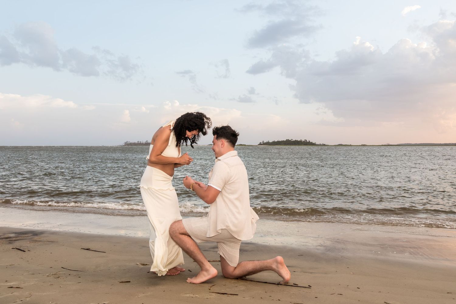 Man proposing on one knee to woman in white dress on sandy beach at sunset near calm water.