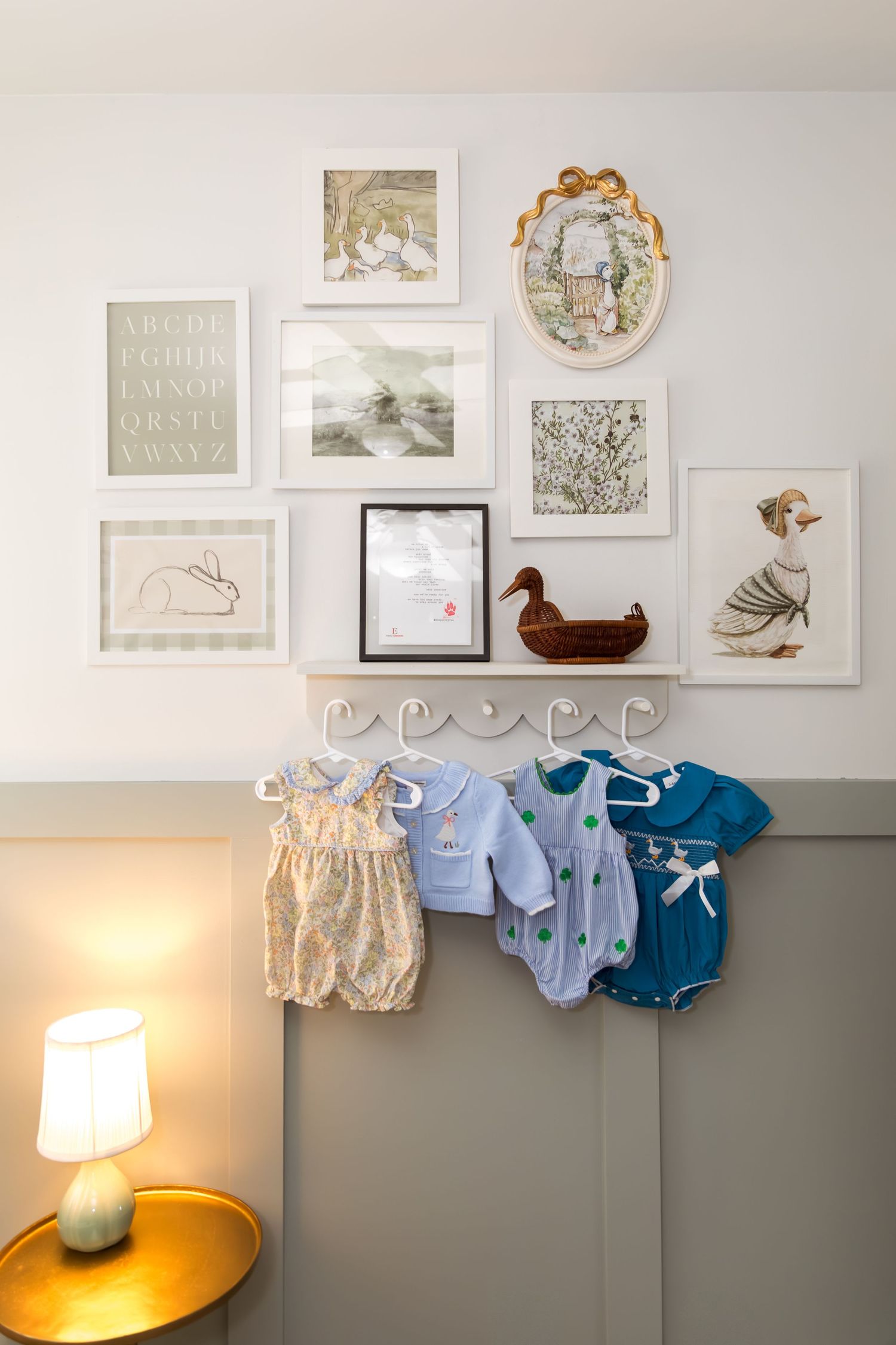 Baby nursery wall with framed art prints, duck decor, and colorful infant onesies hanging on white hooks above a glowing lamp.
