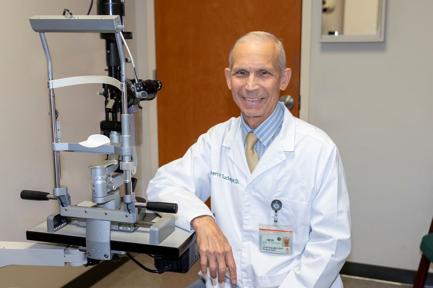 A smiling male doctor in a white lab coat poses next to an ophthalmology slit lamp in a medical examination room.