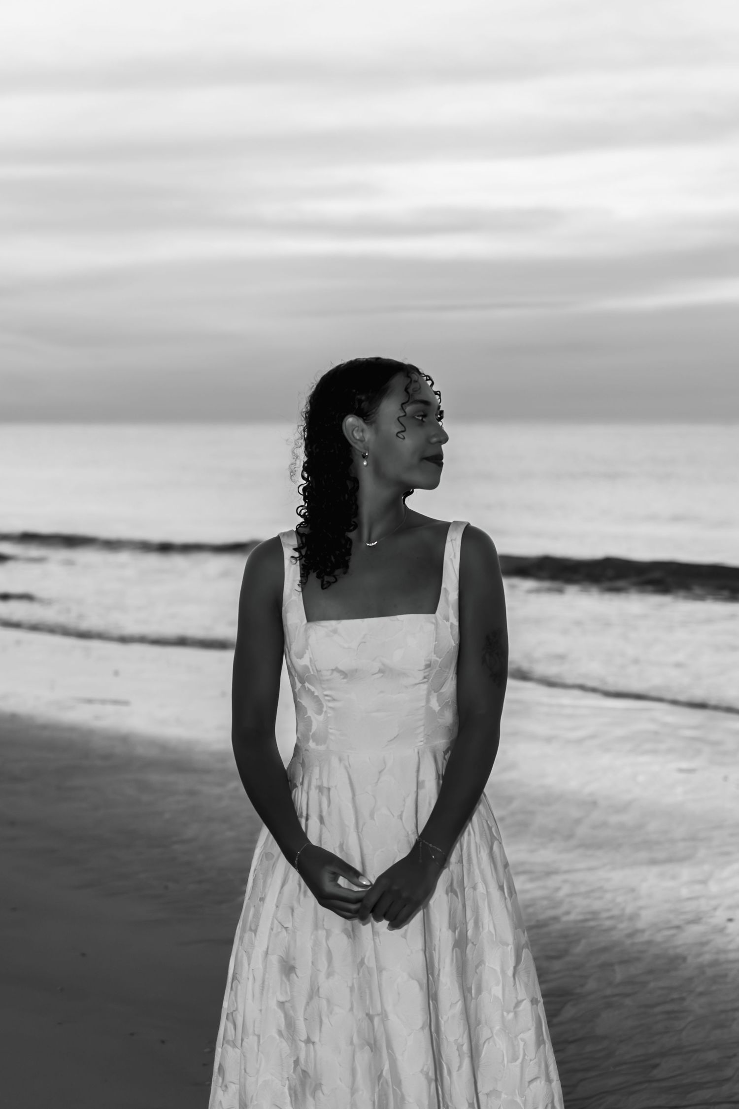Black and white photo of a woman in a white dress standing on a beach at dusk, gazing into the distance.