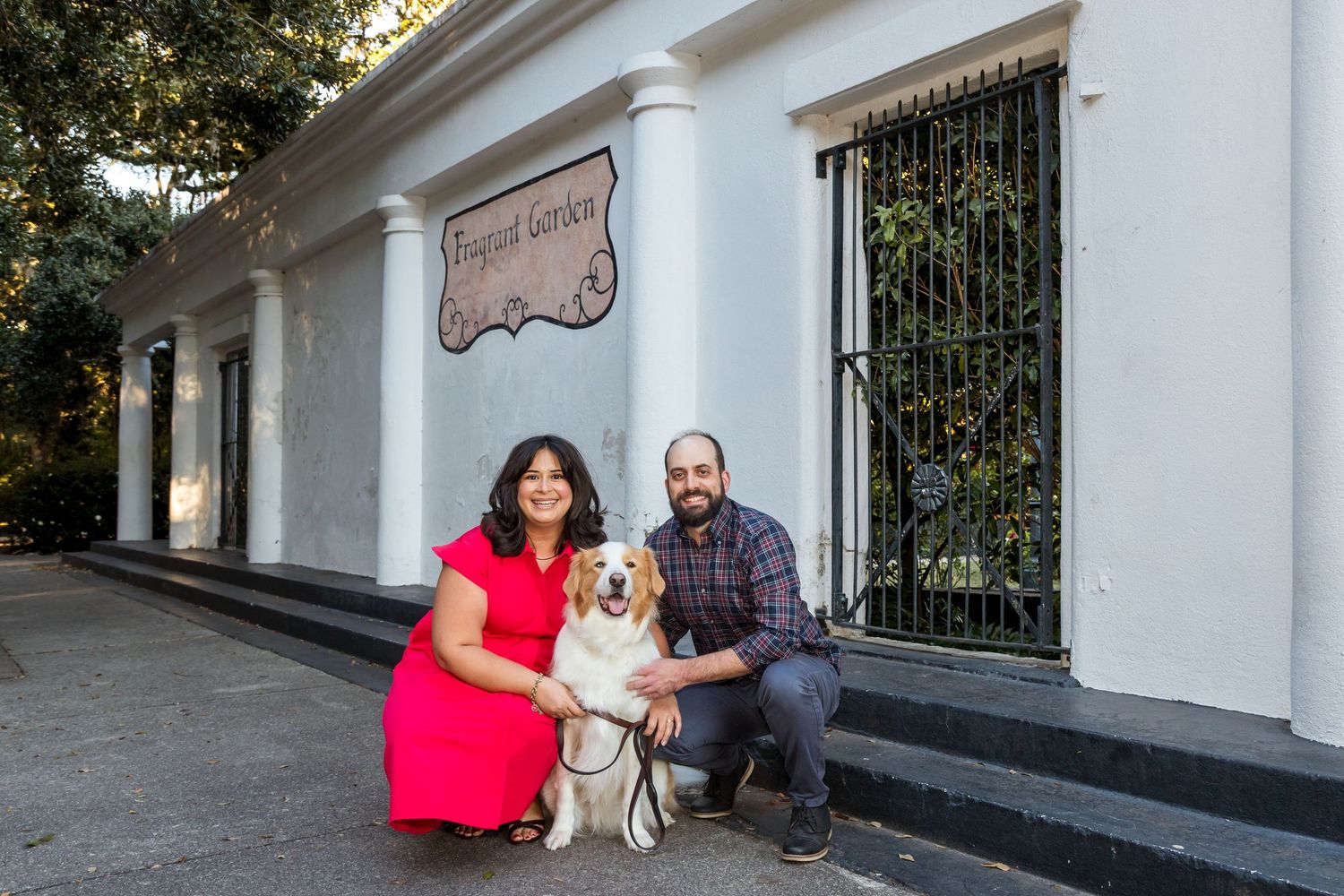 Couple posing with golden retriever dog in front of white building with decorative windows.
