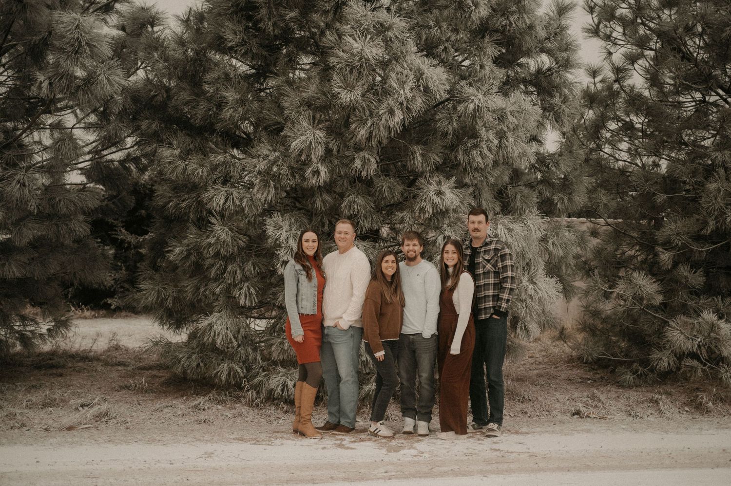 A group of people standing together outdoors in front of evergreen trees in a moody sepia-toned photograph.