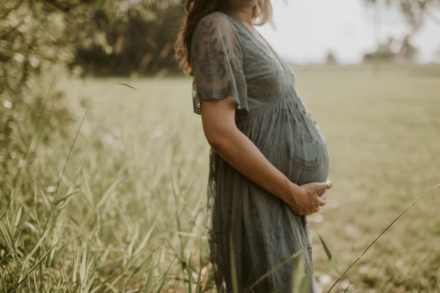 A pregnant woman in a flowy gray dress stands in a sunlit meadow during a maternity photoshoot.