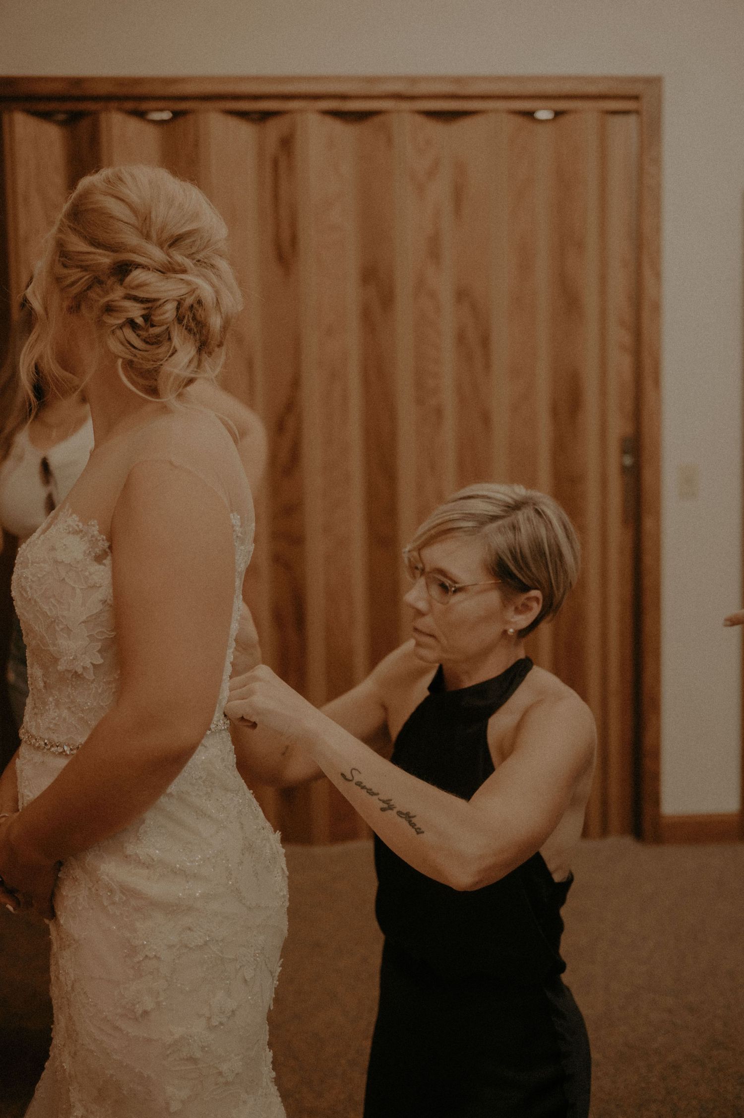 A person in a black dress helps adjust a white lace wedding gown in a warm-toned indoor setting.