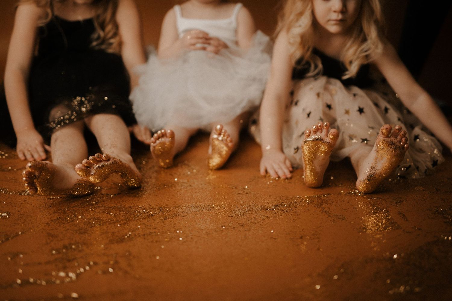 Multiple pairs of bare feet stand on a warm brown surface with scattered glitter creating a magical atmosphere.