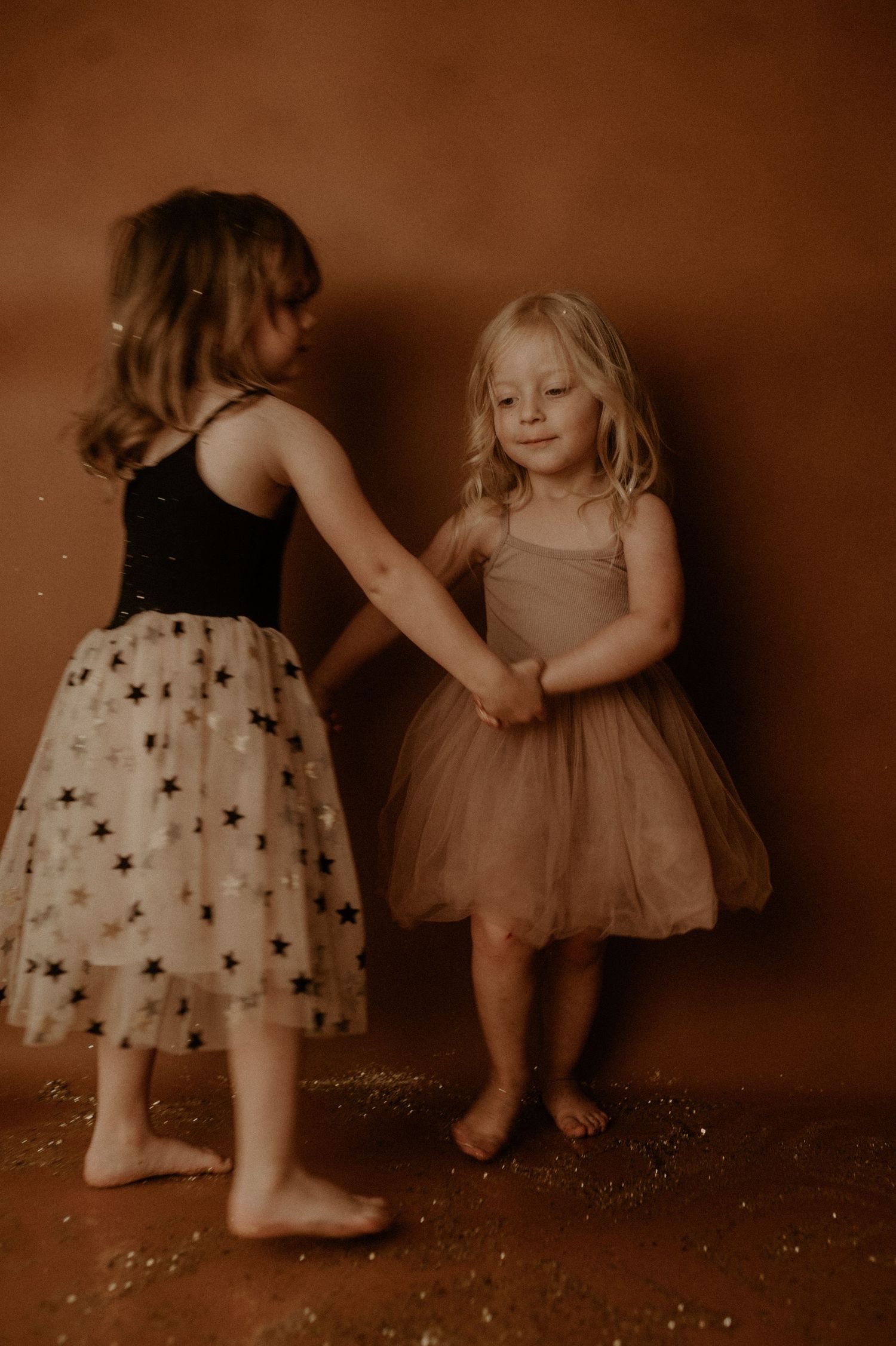 Two small children in tulle dresses play together in a warm sepia-toned photo series against a dark background.