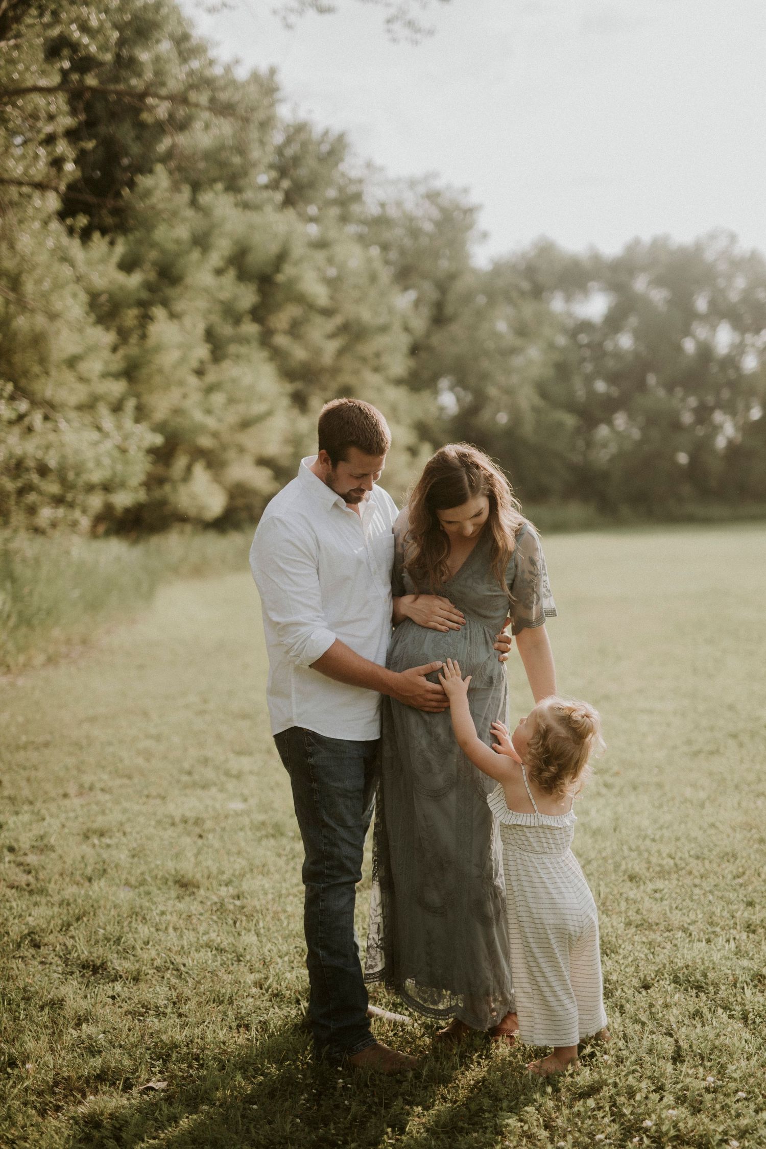 A family shares a tender moment together in a sunlit meadow, dressed in white and gray tones.