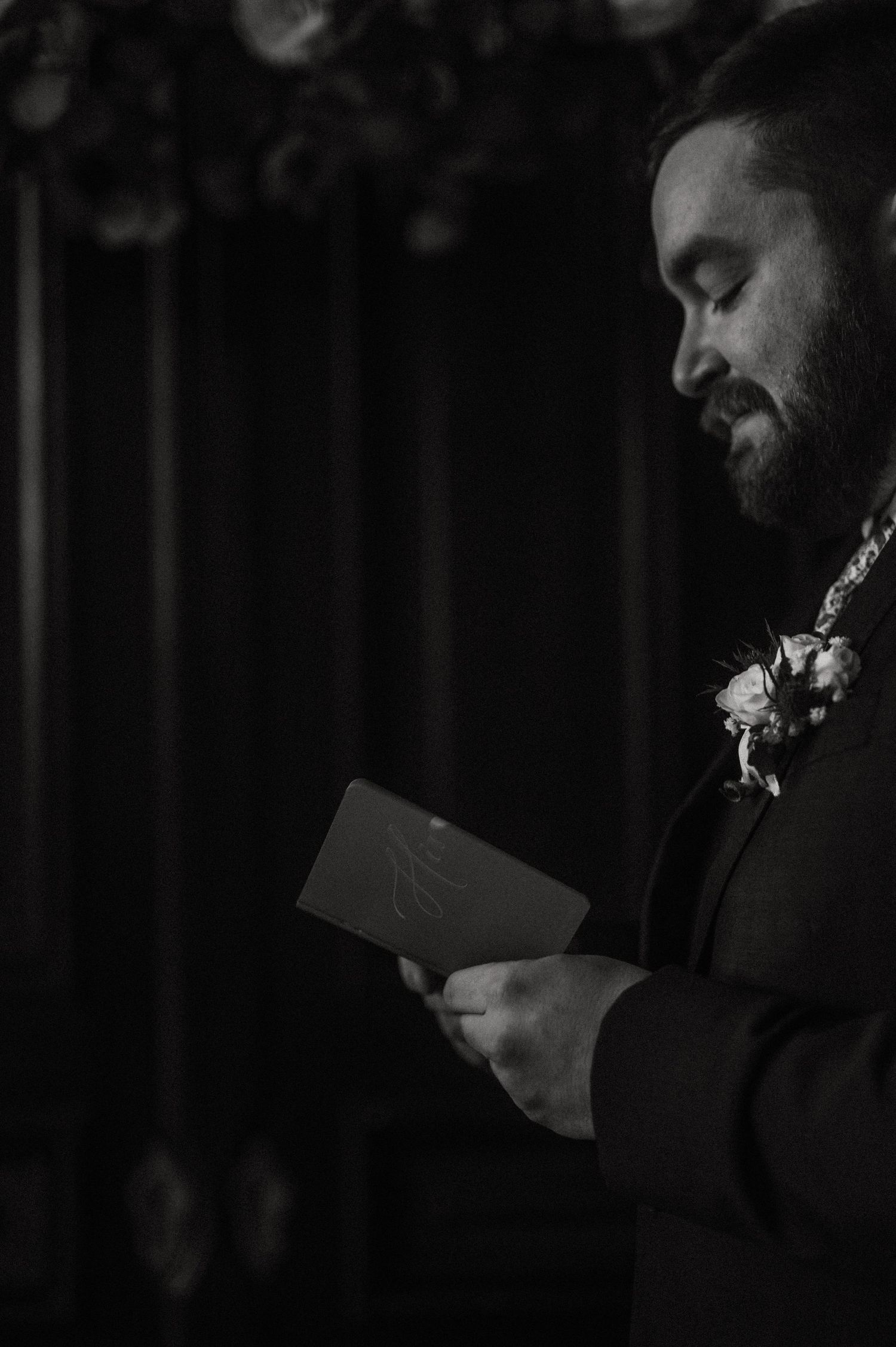 A black and white portrait of someone reading from a small card during a ceremony.