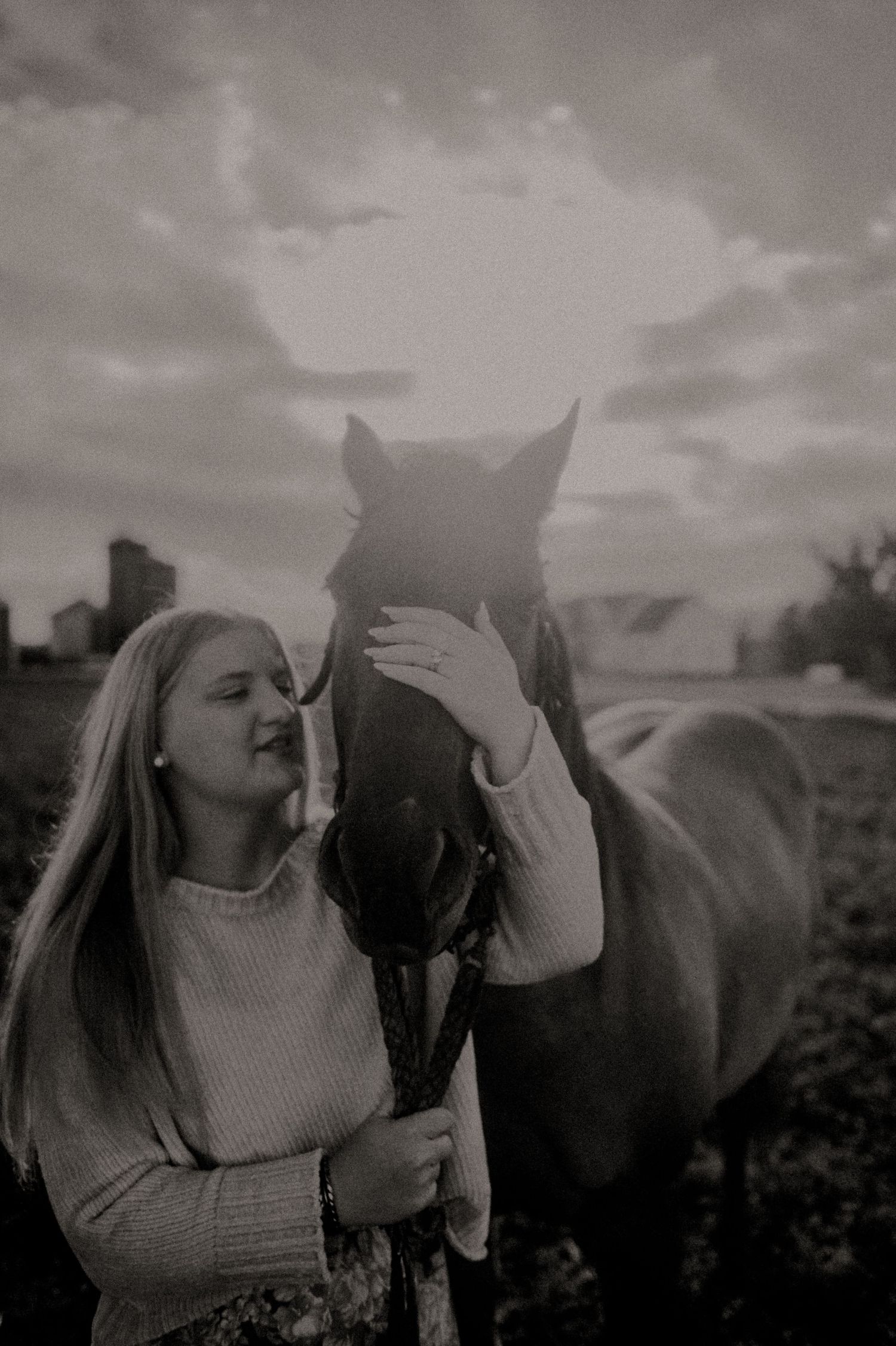 Woman in white sweater lovingly pets a dark horse's face against a cloudy sky.