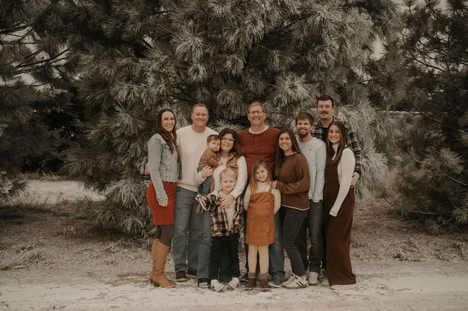 A multi-generational group photo taken outdoors with pine trees in the background in warm earth tones.