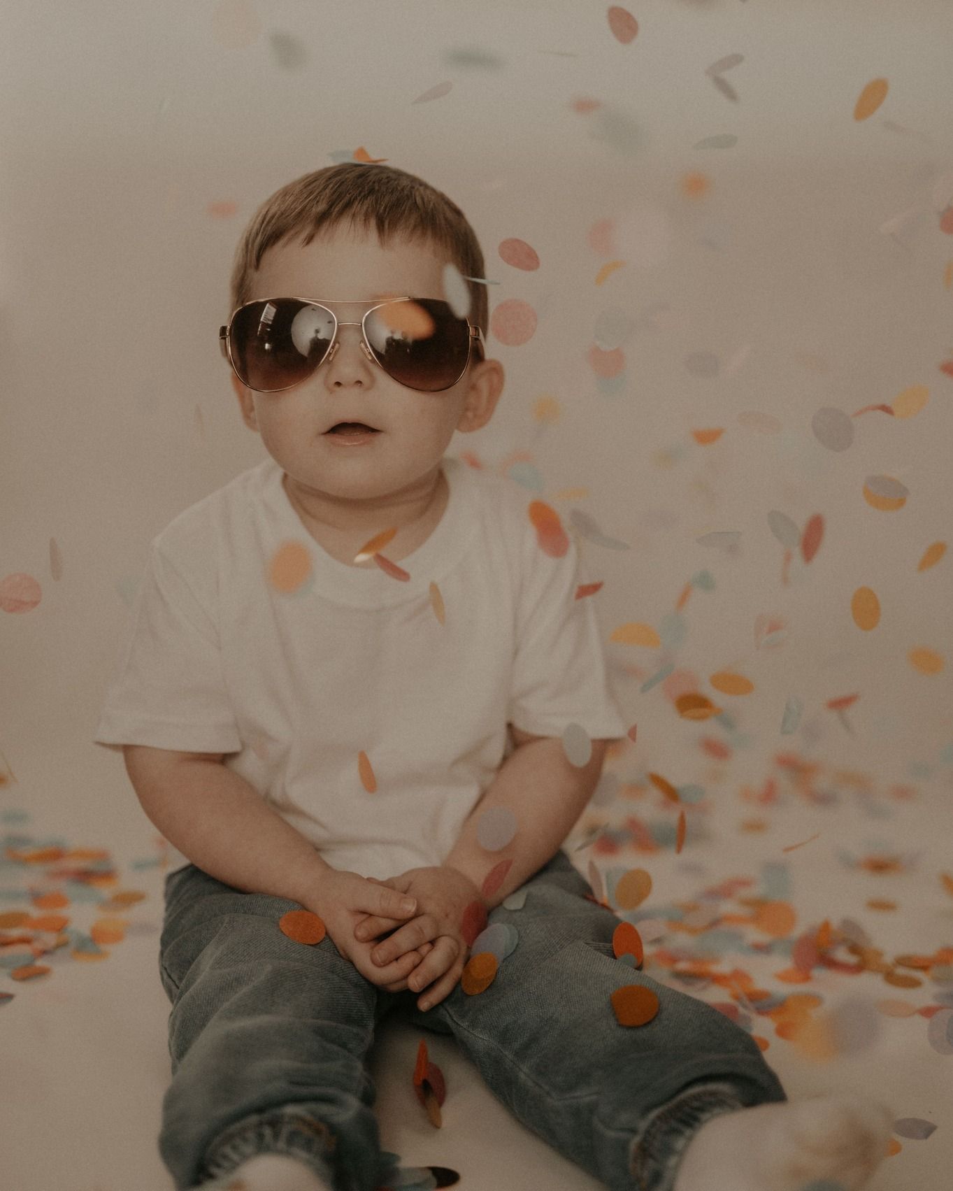 A child wearing sunglasses sits among scattered orange confetti in a playful photo shoot.