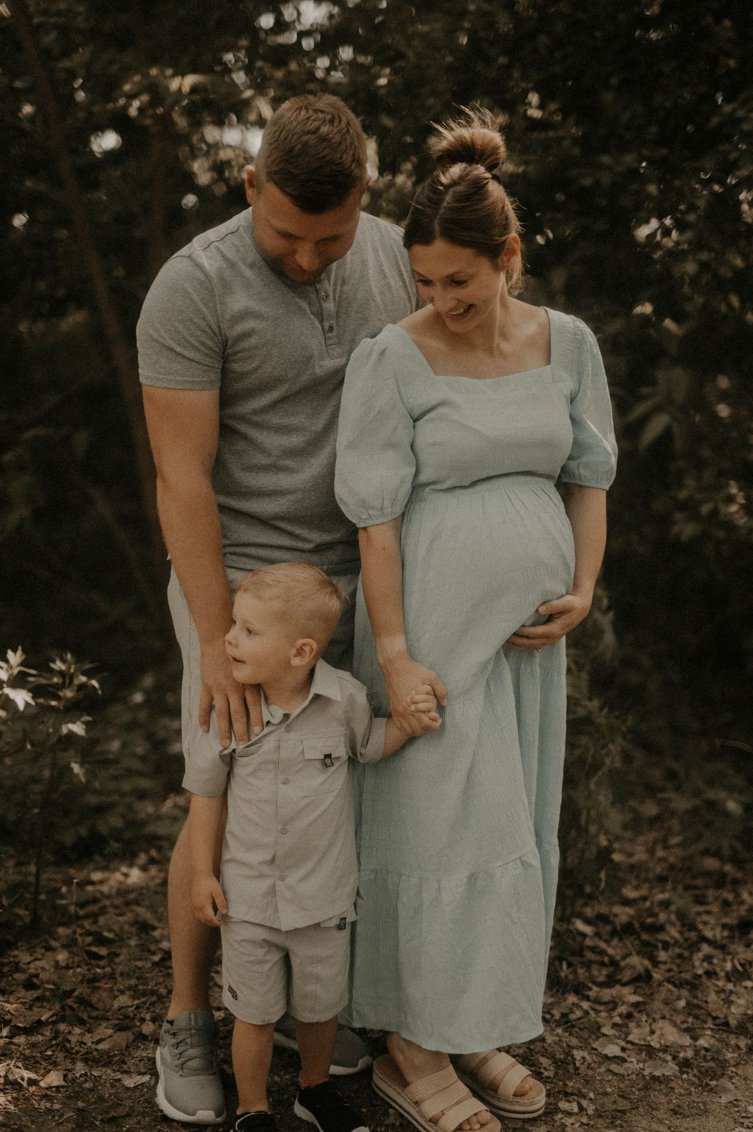 Family in coordinated neutral outfits stands together in a wooded outdoor setting during golden hour.