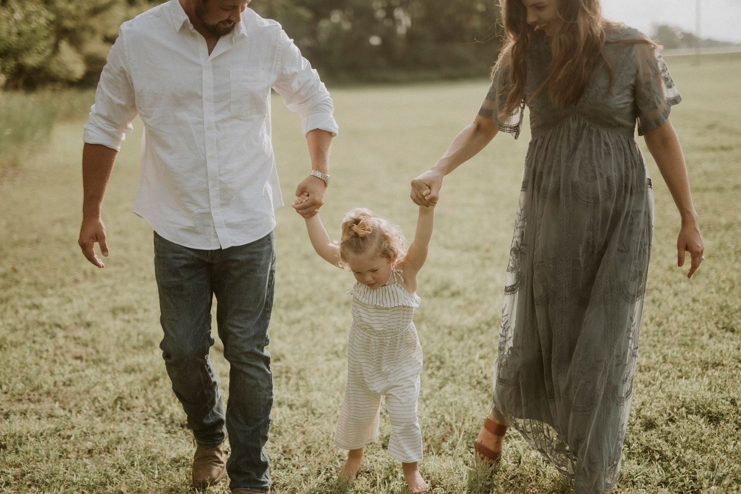 Couple holding hands while walking through a sunny meadow wearing casual white and gray outfits.