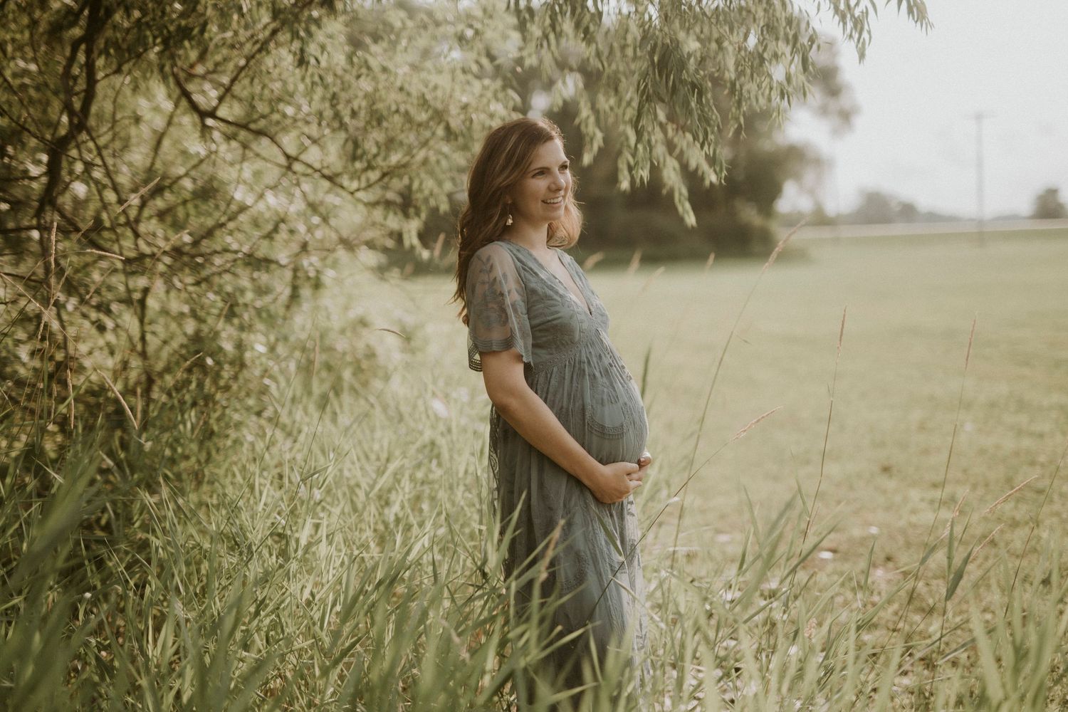 A pregnant woman in a gray dress stands in a sunlit field of tall grass during golden hour.