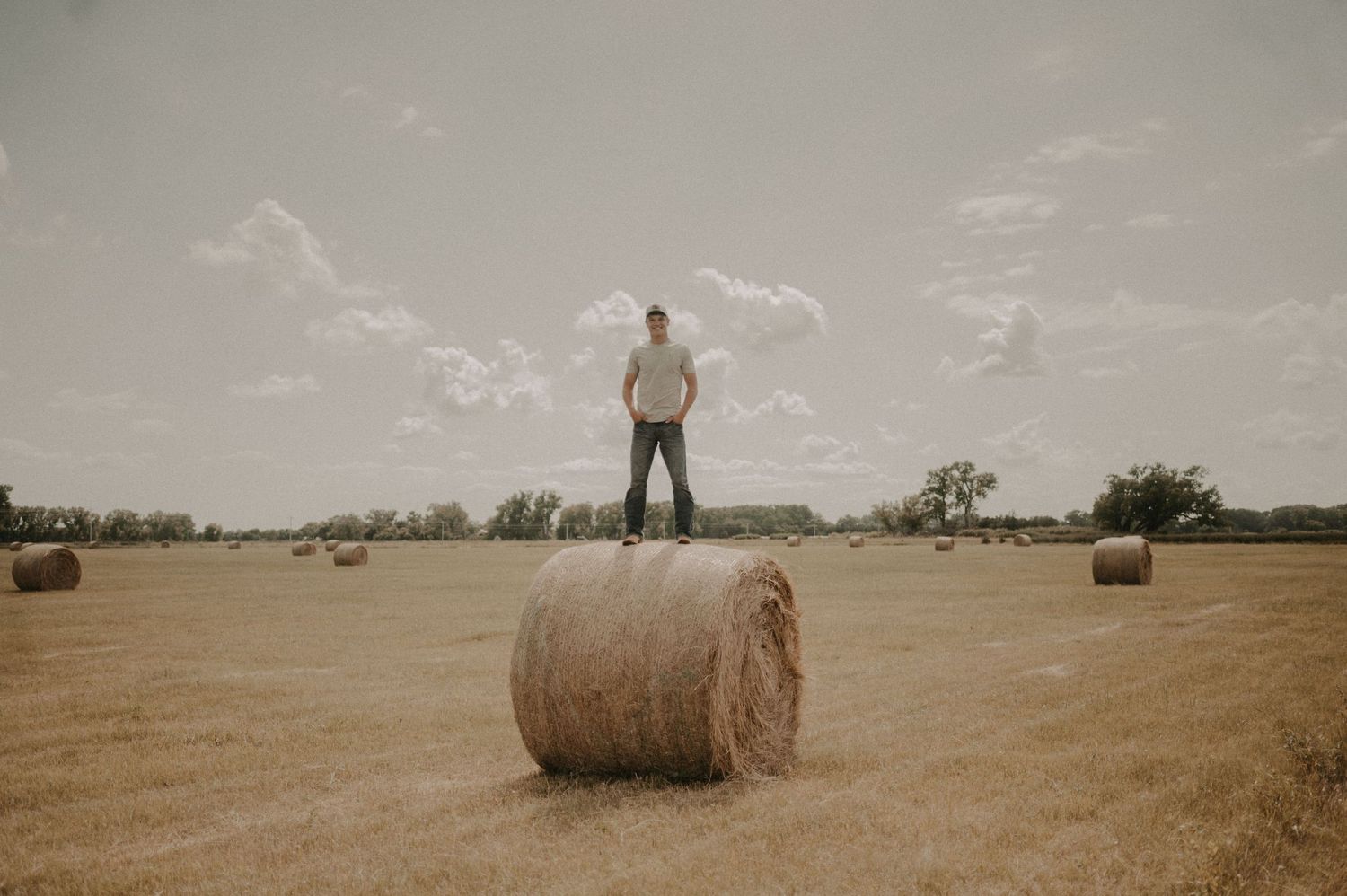Round hay bales dot a golden harvested field under cloudy skies in a rural farming landscape.