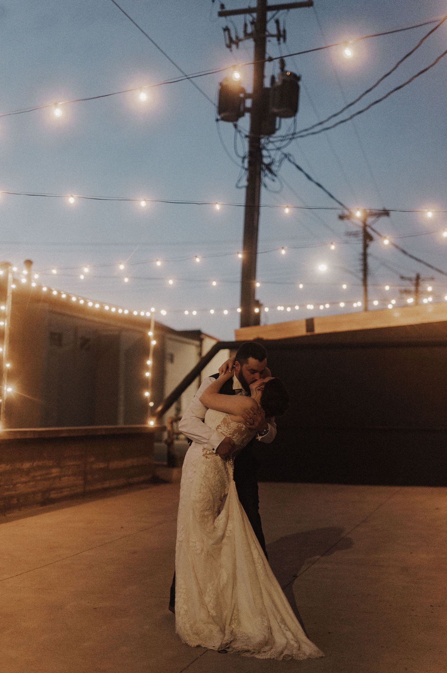 Newlyweds dancing romantically under string lights at dusk with urban backdrop.