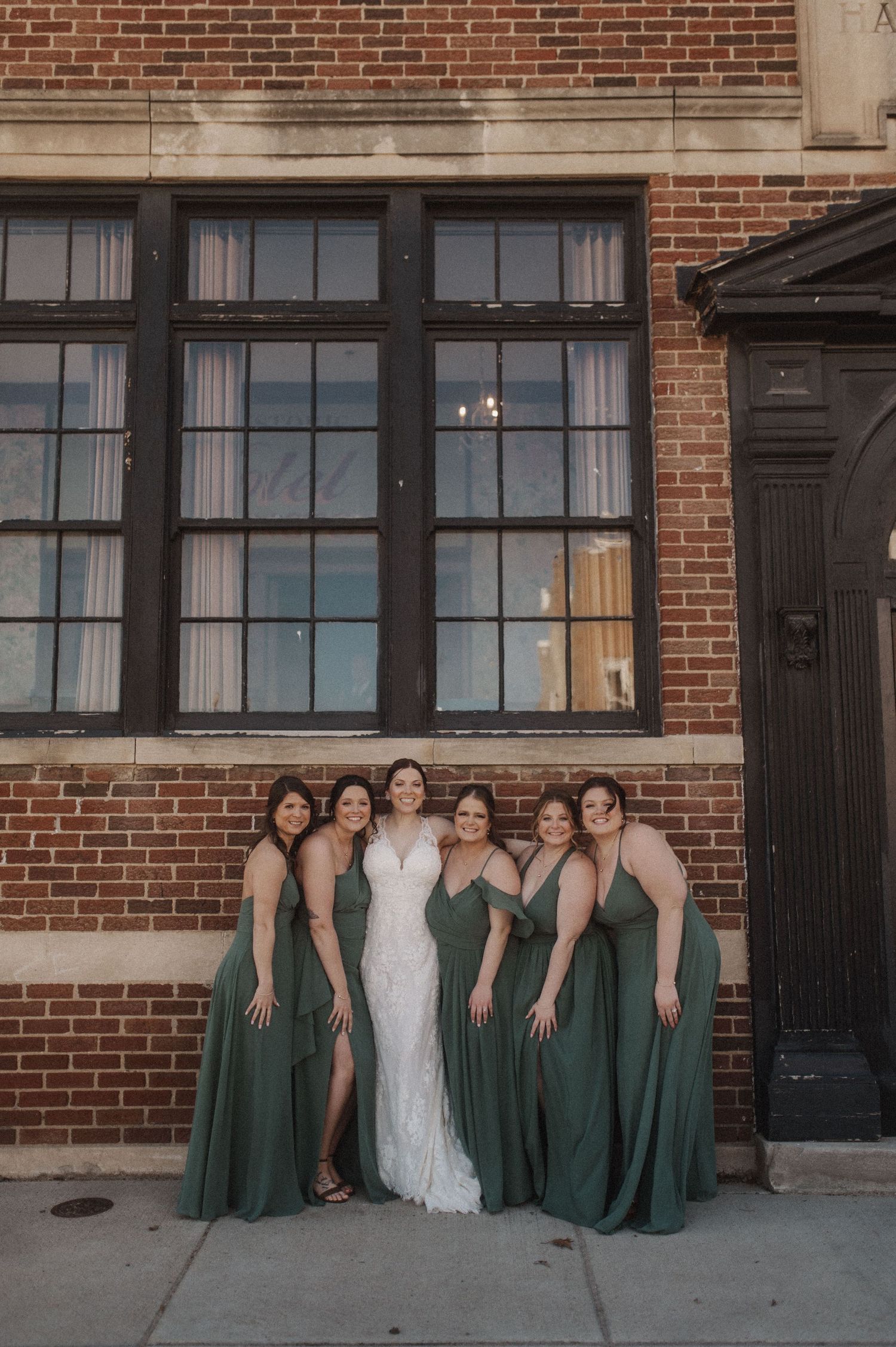 Wedding party in emerald green dresses poses against brick wall exterior.