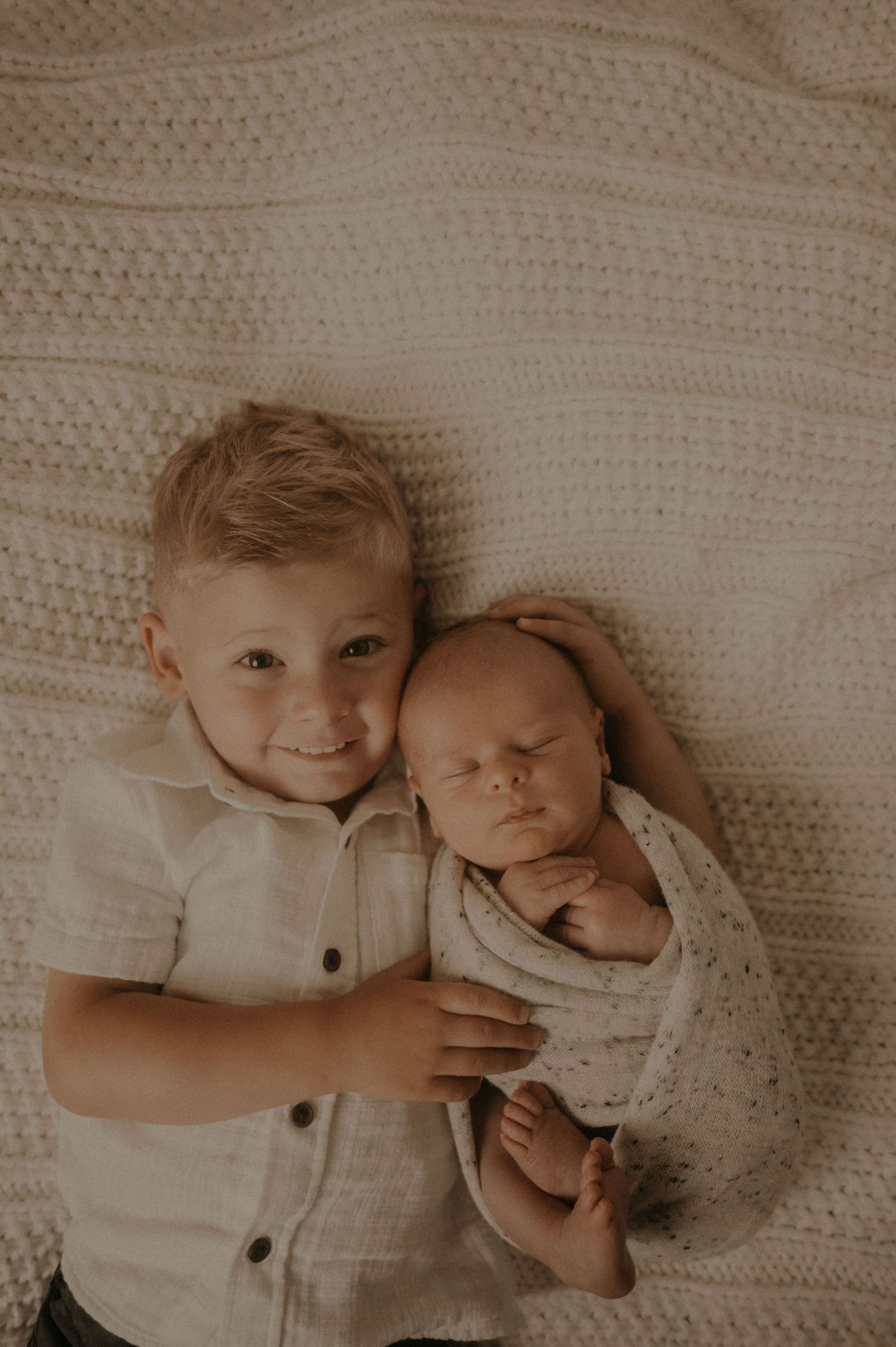 Two siblings in white clothing resting together on a knitted blanket in soft lighting.