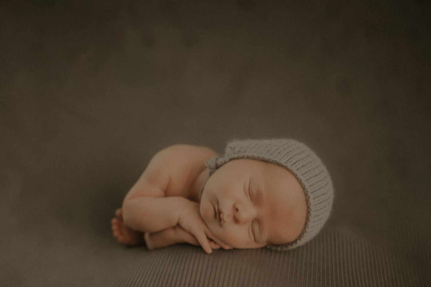 Peaceful newborn baby in fetal position wearing knit hat on textured gray backdrop.