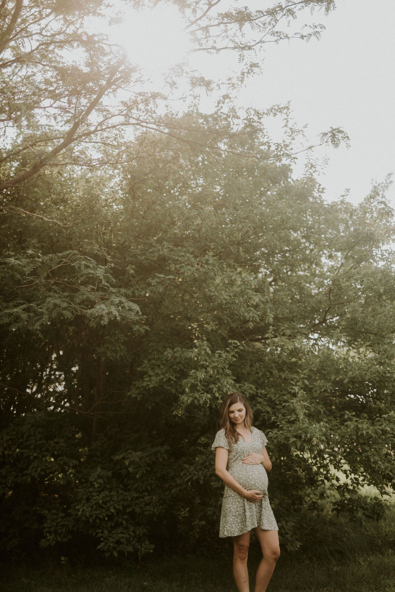 Expecting mother in a floral dress poses outdoors during a golden hour maternity photoshoot in a natural setting.